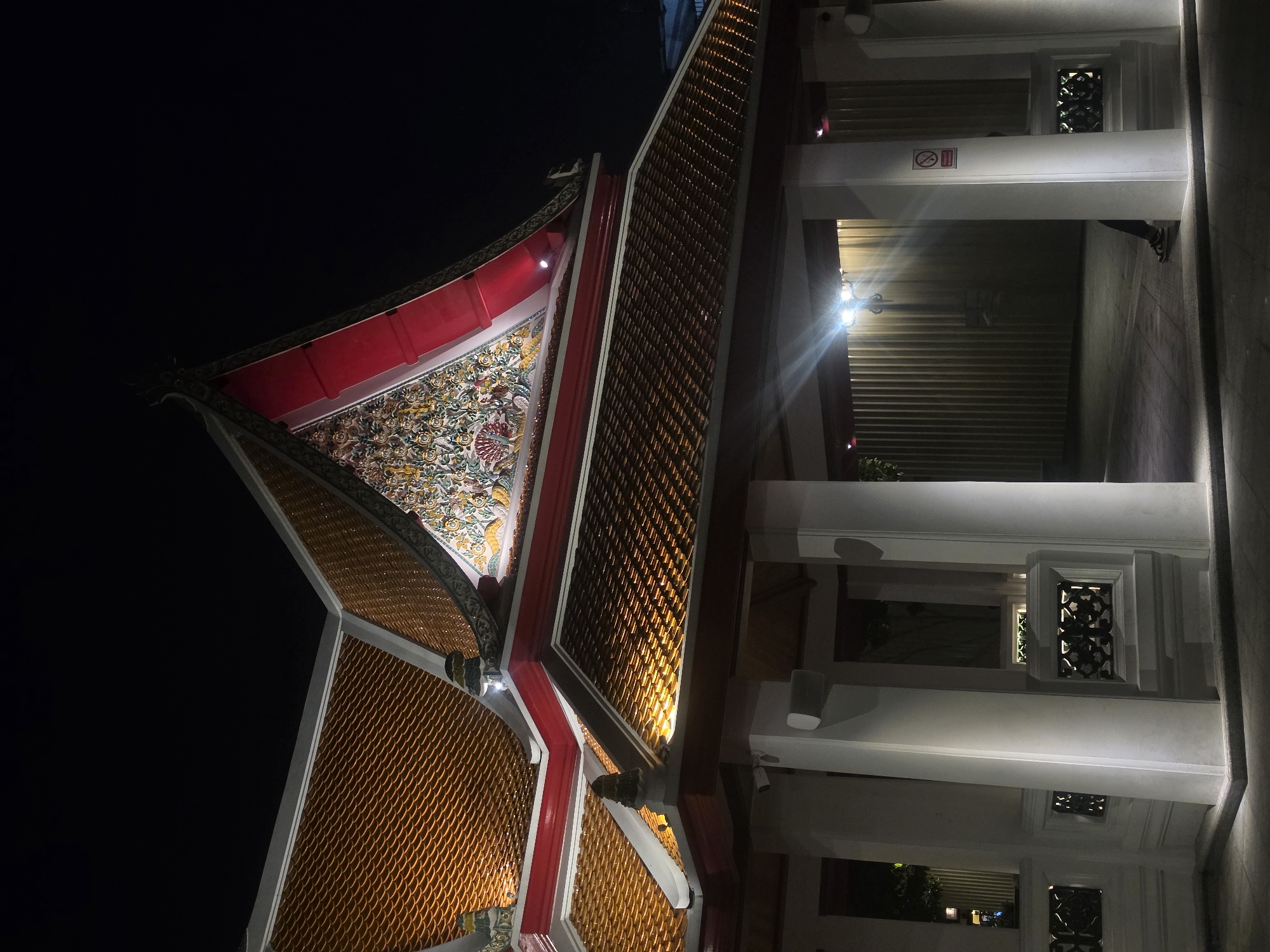 The image shows a nighttime view of architecture in Bangkok, Thailand. The scene includes part of a traditional Thai building with elaborate roof details: layered orange and gold tiles, ornate carvings, and a vibrant mural under a red roof panel. Next to it is a building with white columns and latticed windows. The scene is illuminated by artificial lights.