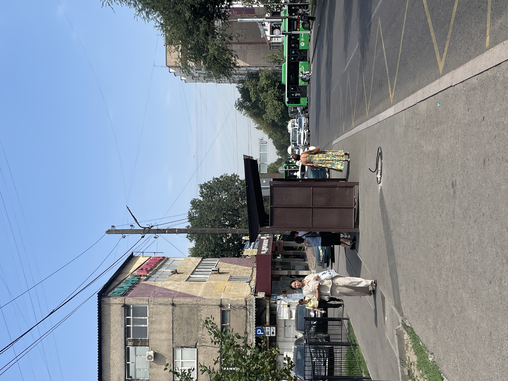 The image depicts a street scene in Almaty, Kazakhstan, featuring a busy road with several green buses and cars. On the sidewalk, a woman in a light-colored outfit walks towards the viewer, carrying a white bag. Another woman in a colorful dress is also visible on the sidewalk, walking away from the viewer. A bus stop shelter is present on the left side of the image. The buildings lining the street show a mix of architectural styles. The sky is clear and blue, indicating a sunny day. The overall scene suggests an urban environment with active pedestrian and vehicular traffic.