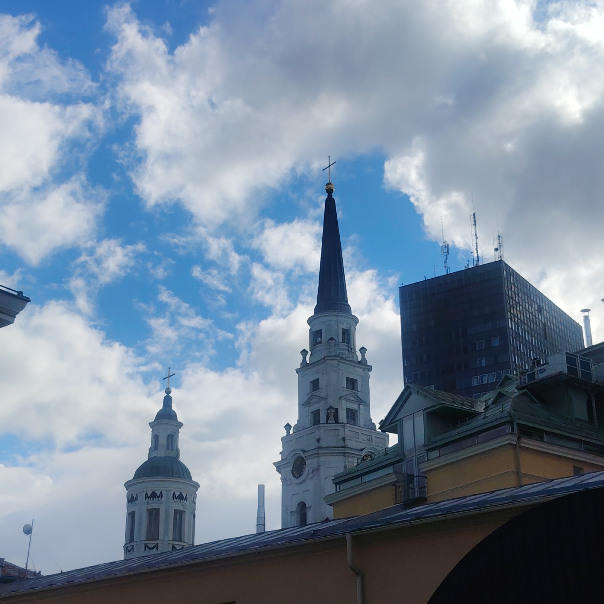 The image captures a scene in Riga, Latvia, showcasing a blend of architectural styles against a sky of scattered clouds. Two church spires dominate the view, one with a classic dome and the other a taller, needle-like spire topped with a cross. Adjacent to these historical structures is a modern, block-shaped building with antennas on its roof. The foreground reveals parts of rooftops with varied designs and colors, including copper roofing, yellow and green facades. No people are visible in the frame.