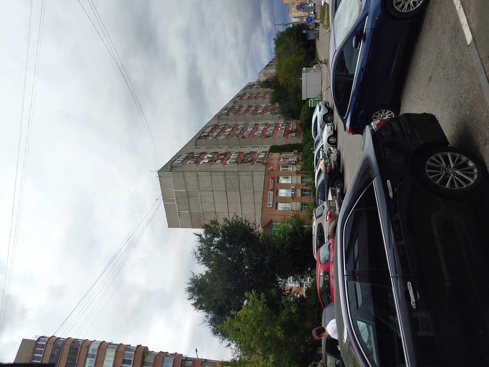 The image depicts a street scene in the city of Zhukovsky, Russia, on a cloudy day. In the foreground, a dark sedan is parked. Behind it, a blue sedan is visible. Further back, a row of parked cars includes a silver sedan, a red hatchback, and a light-colored SUV. A person in a white shirt is standing near the driver's side of a black car.
Dominating the background are two large residential buildings. The building on the left is a tall, multi-story structure with a curved facade and numerous windows. To its right is a longer, concrete building with a grid-like pattern of windows. At the base of the concrete building, there is a smaller, brick building with a sign reading "АЛЬТАИР Медицинский центр" (ALTAIR Medical Center). Trees are interspersed between the buildings and along the street. Overhead, power lines stretch across the sky. In the distance, more buildings and a street with a bicycle lane and pedestrians are visible.