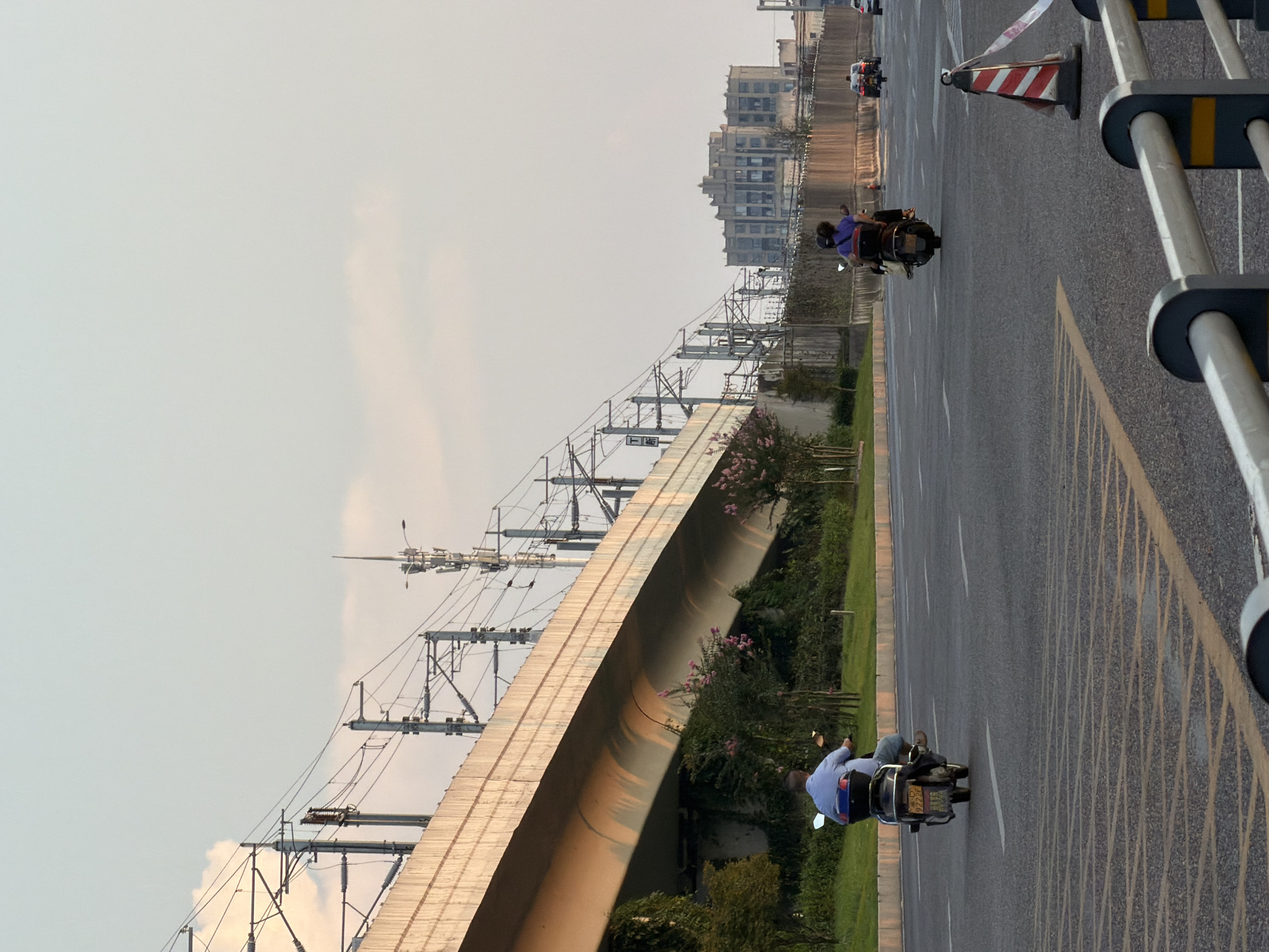The image captures a street scene in Jiangjin, China, under a pale sky with sparse clouds. A multi-level elevated structure, likely a highway or railway viaduct, dominates the left side of the frame, supported by concrete pillars and featuring overhead power lines and communication towers. Greenery, including trees and bushes with pink flowers, lines the base of the elevated structure.
On the road, two individuals are seen riding scooters. In the foreground left, a person in a light blue shirt and blue jeans is riding a scooter with a black storage box. The scooter has a license plate with visible characters. Further down the road, another scooter carries two individuals, one in a purple top. Several cars are visible in the distance, also traveling on the road.
The road itself is asphalt, marked with yellow and white lines, and features a textured section with yellow markings in the foreground. A red and white striped traffic cone with white tape is positioned on the right side of the road, near a metal barrier. In the background, a large, multi-story building with a distinctive architectural style is visible, suggesting an urban or suburban setting. The overall lighting suggests it is late afternoon or early evening, with warm tones cast by the setting sun on the elevated structure.