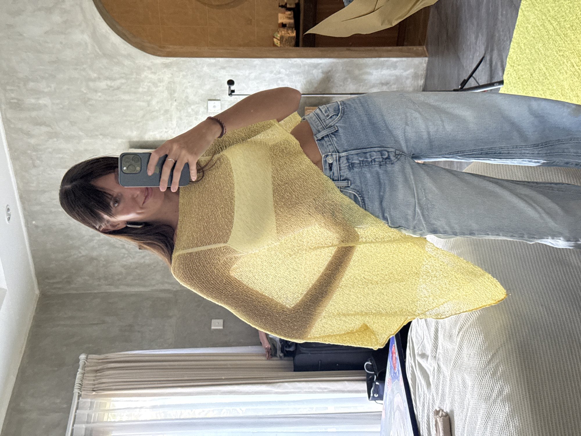 A young woman is captured in a mirror selfie, standing in an indoor room. The image is oriented sideways, with the woman appearing to stand horizontally. She holds a dark smartphone, likely an iPhone, in her right hand, positioned to capture her reflection. Her dark hair, styled with bangs, frames her face as she looks directly at the camera with a neutral expression.
She is wearing a sheer, loosely woven yellow top over a lighter undergarment, possibly a bralette or camisole. This is paired with light-wash blue jeans. A braided bracelet adorns her right wrist, and a ring is visible on her right hand's ring finger.
The background, seen in the mirror's reflection, reveals a room with concrete-textured walls, suggesting a modern aesthetic. An arched wooden doorway or decorative element is visible in the upper part of the reflection. To the left, a window with beige-colored blinds or curtains allows natural daylight into the room. A bed with white bedding is partially visible on the right, and a black suitcase or bag rests on the floor below the window. Another light-colored textured surface, possibly a blanket or rug, is also present. The overall setting appears to be a bedroom or hotel room, consistent with the provided location context of North Kuta, Indonesia. There is no visible text in the image.