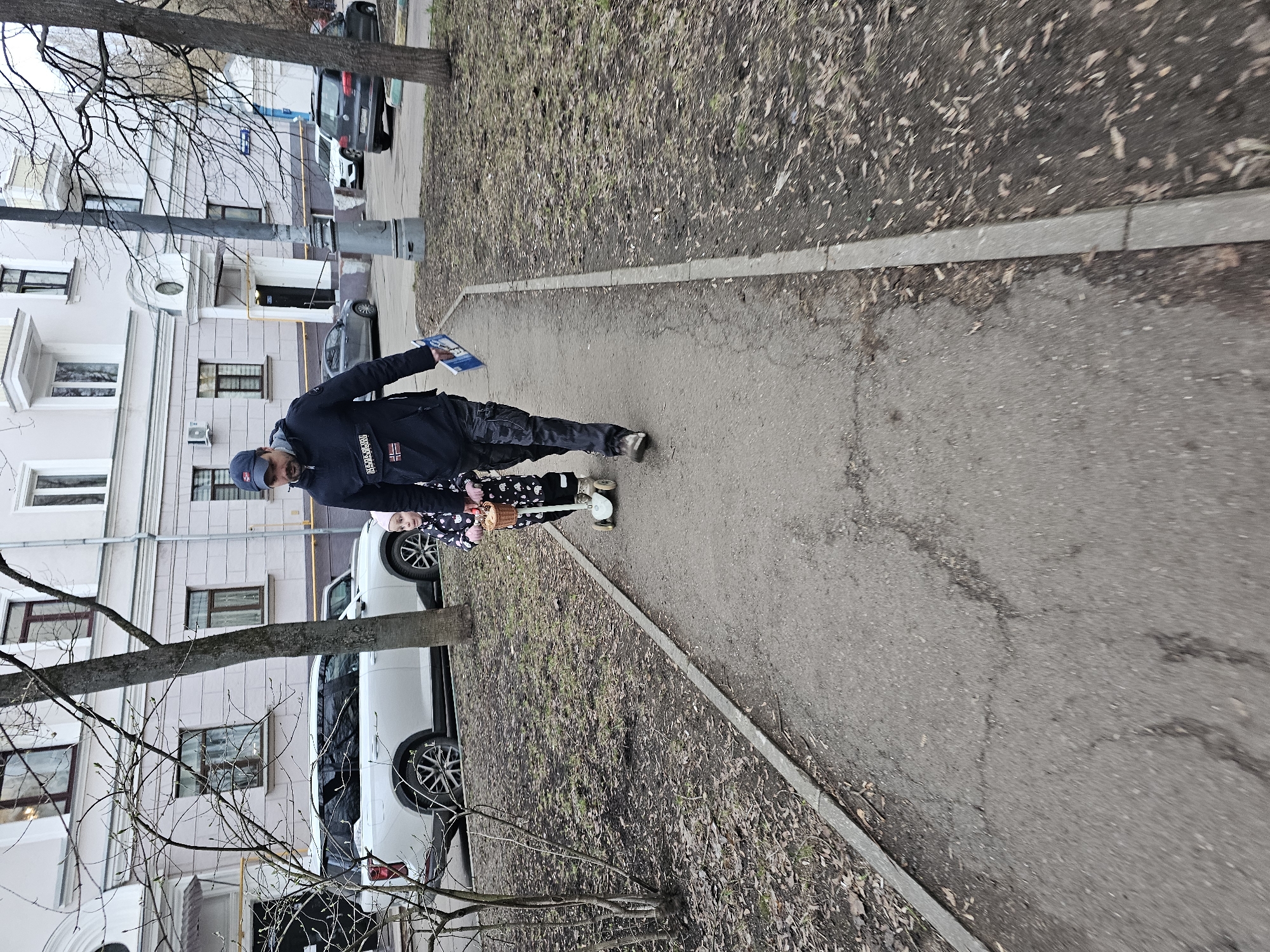 A medium-shot photograph captures a man and a child on a paved pathway beside a grassy area in Khimki, Russia. The man, wearing a dark jacket with a prominent "Napapijri" patch, dark pants, and a baseball cap, is holding a book or magazine. The child, dressed in black and white, is riding a scooter with a basket attached. The pathway is edged with a concrete curb, and dry leaves are scattered on the grass. In the background, a light-colored building with multiple windows and parked cars are visible, suggesting an urban residential area.