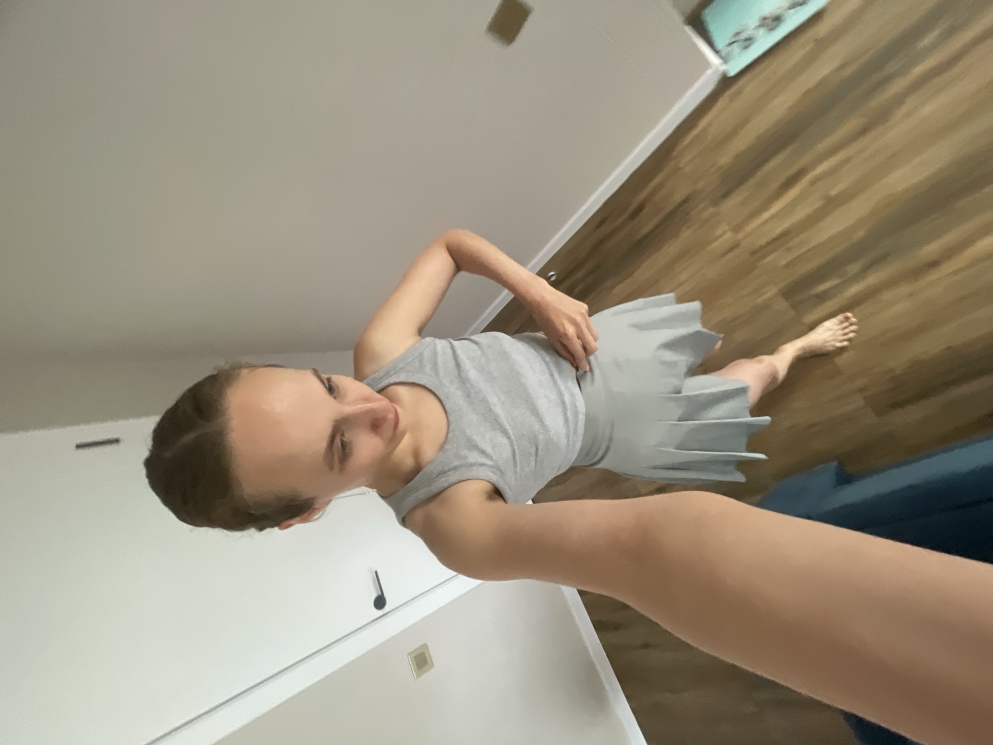 A person is taking a selfie in a room. The person is wearing a grey tank top and a grey pleated skirt. They are standing on a wooden floor. A blue couch is visible in the background. The room appears to be a domestic setting, possibly a bedroom or living area, with white walls and doors. The lighting suggests it is daytime. The person's expression is neutral. There is no visible text in the image. The location context provided is Подшивалово, Russia.