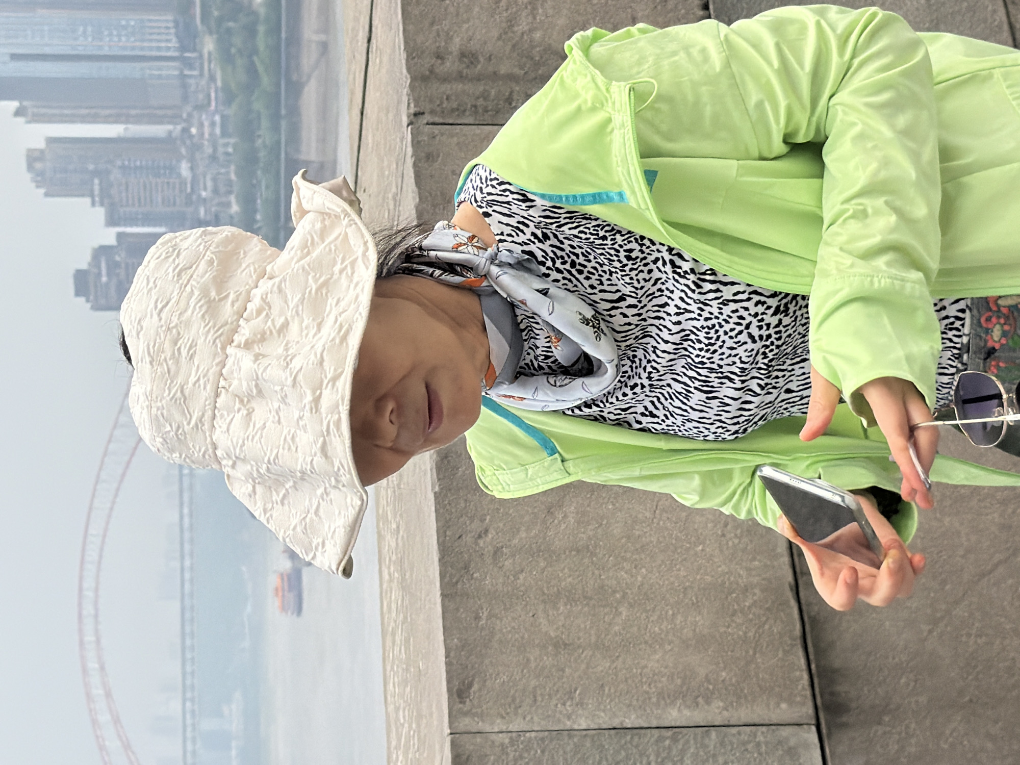 A woman wearing a wide-brimmed hat, a light green jacket, and a patterned shirt stands on a raised walkway with a city skyline and river in the background. The woman is holding a smartphone in her left hand and sunglasses in her right hand. The city skyline features tall buildings and a bridge crossing a river. The woman's face is partially obscured by her hat, but she appears to be looking down at her phone. The scene is likely in Yuzhong District, China, given the urban landscape and architectural style.