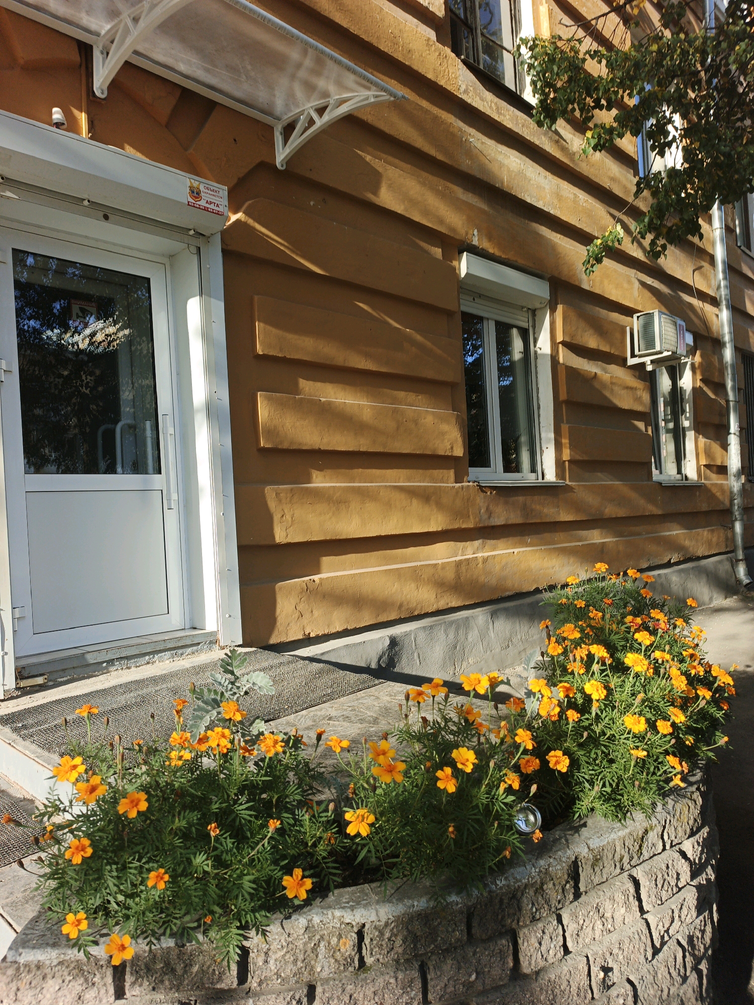 The image shows a building with a yellow wooden facade, a white door, and a window with a rolling shutter. Orange marigold flowers are planted in a stone planter in front of the building. An air conditioning unit is mounted on the wall above one of the windows. The building appears to be located in Zlatoust, Russia, as indicated by the sign on the door which reads "ОБЪЕКТ ОХРАНЯЕТСЯ" and "АРТА", common for security companies in Russia. The scene is bathed in sunlight, casting shadows on the building. There are no people visible in the image.