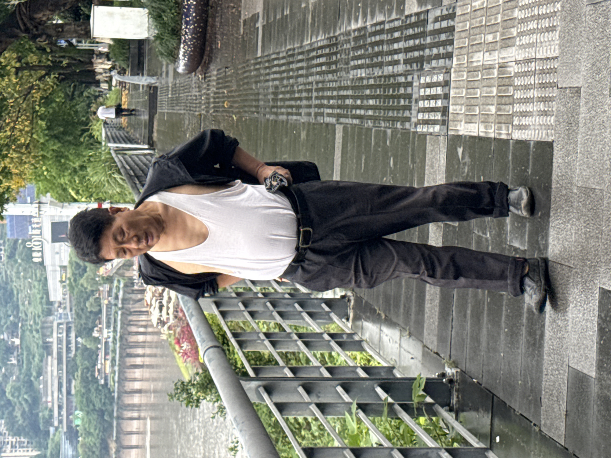A middle-aged man in a white tank top and black pants stands on a grey tile sidewalk, leaning against a grey metal railing. He wears a black jacket draped over one shoulder and black shoes. The man looks down. Behind the railing is a river, lush green vegetation, and buildings, indicating an urban area. In the background are other figures walking along the path. The location is consistent with a riverside promenade in the Nan'an District, China. The weather appears overcast and possibly damp based on the sheen of the sidewalk tiles.