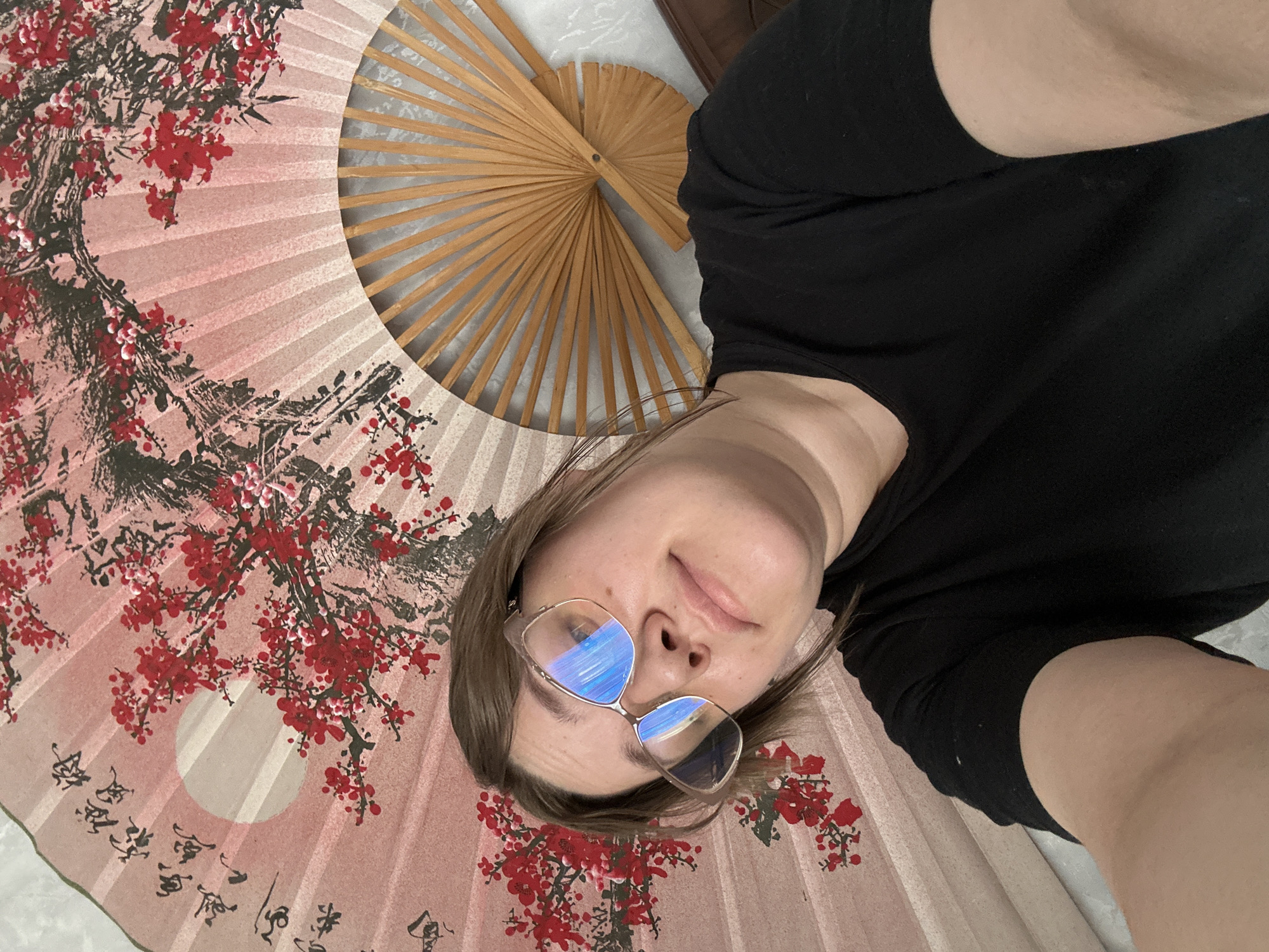 An overhead selfie captures a person, likely female, lying on a large decorative fan with an East Asian design. The subject wears a black top and glasses that reflect blue light, displaying a neutral to slightly smiling expression. The prominent fan serving as the backdrop features a pale pink background adorned with dark, gnarled branches and vibrant red blossoms, reminiscent of cherry or plum flowers. A distinct white circular shape, possibly representing the sun or moon, is visible on the fan's surface, alongside delicate black calligraphic script. To the upper right of the frame, part of a smaller, open wooden fan with visible bamboo ribs is also present. The scene depicts an indoor setting. This media file originates from Ольховка, Russia.