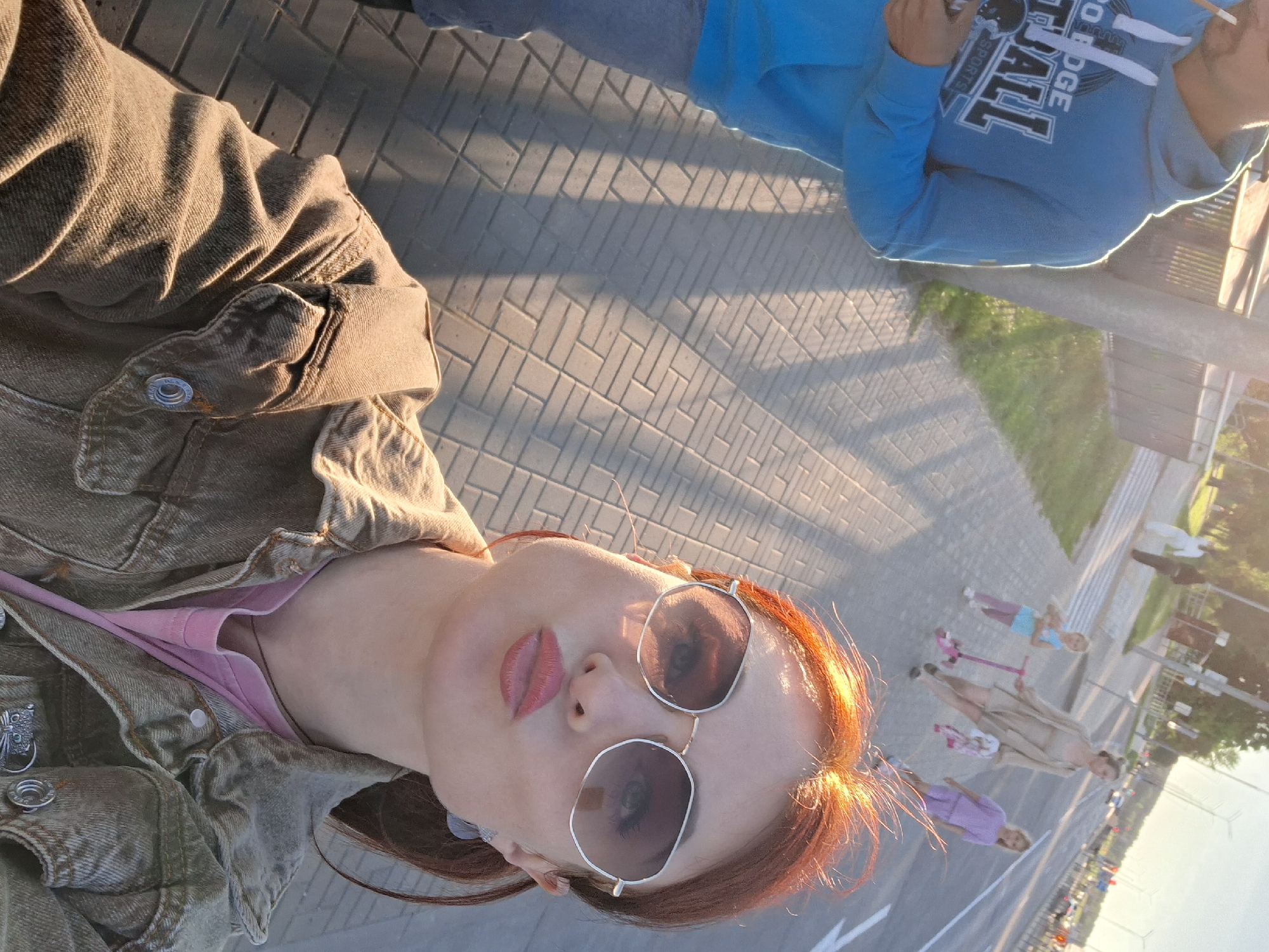 An outdoor, eye-level, medium shot captures a selfie taken by a woman with red hair and oversized sunglasses. She has her cheeks puffed out and is wearing a light brown denim jacket over a pink t-shirt. Her right arm, holding a smartphone, is visible in the lower left corner. In the background, a man in a blue hoodie walks away from the camera. Further in the distance, a woman pushes a stroller with a child on a scooter, followed by another child on a scooter. The scene appears to be a paved sidewalk next to a road, likely in Smalyavichy, Belarus, given the presence of streetlights and traffic. The lighting suggests it is late afternoon or early evening.
