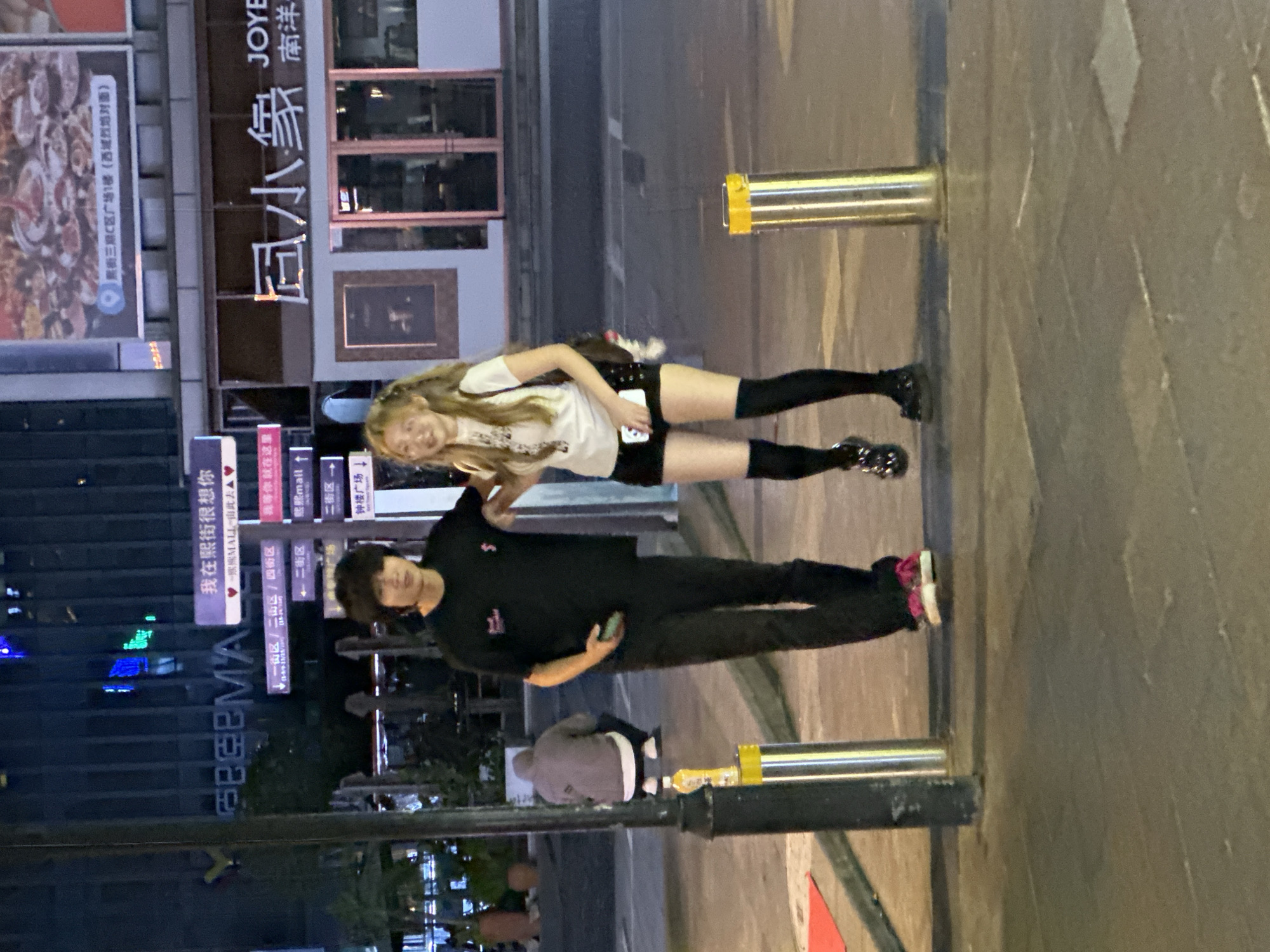 Here is a description of the image:
The image shows two people standing next to each other in front of a building at night. A woman with long blonde hair is wearing a white t-shirt, black shorts, and black knee-high socks. She is standing next to a man wearing a black t-shirt and black pants. The man is holding a cell phone.
The background includes a large sign written in Chinese, likely advertising "Joy City," and text indicating the location is in the city of Chongqing High-tech Zone, China. There is also other signage related to a shopping mall. Bollards and other street fixtures are visible in the foreground.