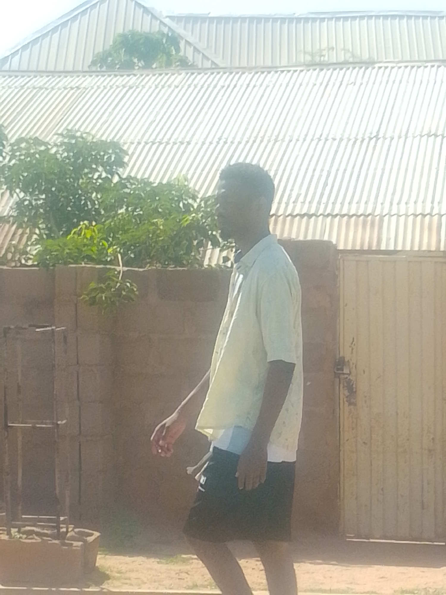 A young Black man walks outdoors in a dusty, sunlit environment. He is wearing a light-colored, short-sleeved button-down shirt over a white undershirt and black shorts. His right arm is bent at the elbow, and his left arm is slightly forward. He is walking from left to right in the frame.
The background features a corrugated metal roof that appears to be part of a building. Some green foliage is visible on top of a low, cinder block wall. To the right, a weathered, yellow metal gate is visible, with a lock on it. The ground is sandy and dry, with some sparse grass. In the lower left, there is a partially constructed metal frame structure.
The lighting suggests it is daytime, likely mid-morning or afternoon, given the bright sunlight and distinct shadows. The overall impression is a casual, everyday scene in a warm climate. There is no visible text in the image.