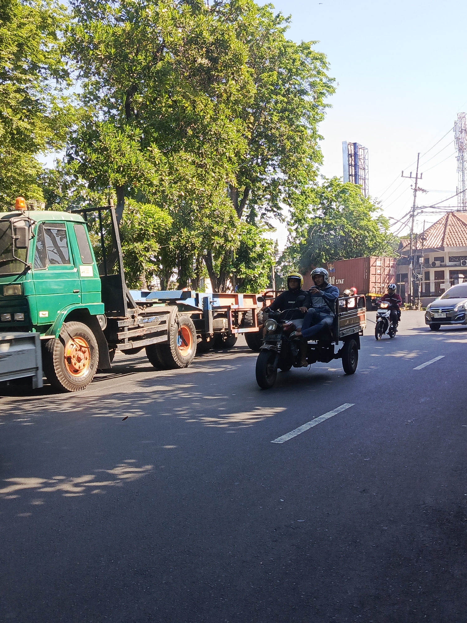 This image depicts a street scene in Surabaya, Indonesia, under a clear sky and bathed in bright sunlight that casts tree shadows across the asphalt. A large green truck with a flatbed trailer is prominently featured on the left side of the frame, its tires showing orange rims. In the middle ground, a three-wheeled motorized vehicle, also known as a "becak motor," is carrying two male passengers, both wearing helmets and casual clothing. The passenger in front is wearing a dark jacket, and the one behind him is wearing a blue jacket. To the right of the becak motor, another motorcycle with a rider wearing a maroon shirt and helmet is moving in the same direction. Further to the right, a silver sedan is visible, and behind the becak motor, a shipping container with the text "LINE" is on a trailer. The background reveals trees, buildings, and utility poles, consistent with an urban environment. The overall impression is a snapshot of daily life and transportation in Surabaya.