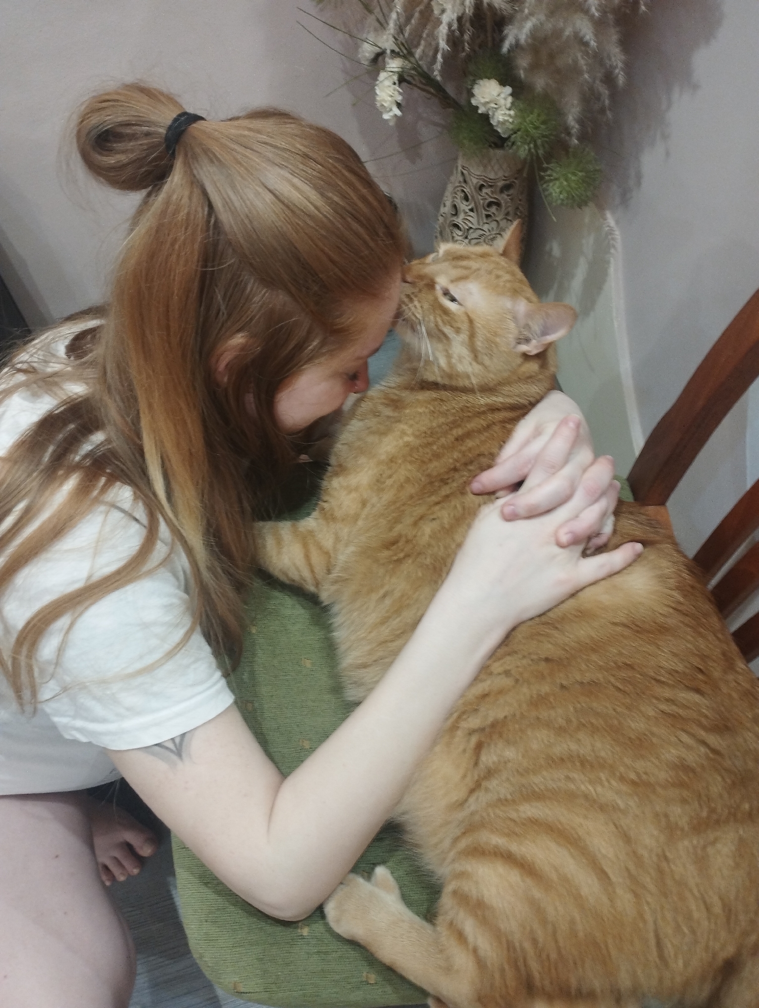 A woman with long, light red hair pulled back in a bun is embracing a large, orange tabby cat. The woman is kneeling on the floor, wearing a white t-shirt. Her arms are wrapped around the cat, and her face is close to its head. The cat is sitting on a chair with a green cushion. Behind the woman and the cat is a wall with a vase containing dried flowers. The location is Unknown, Unknown.