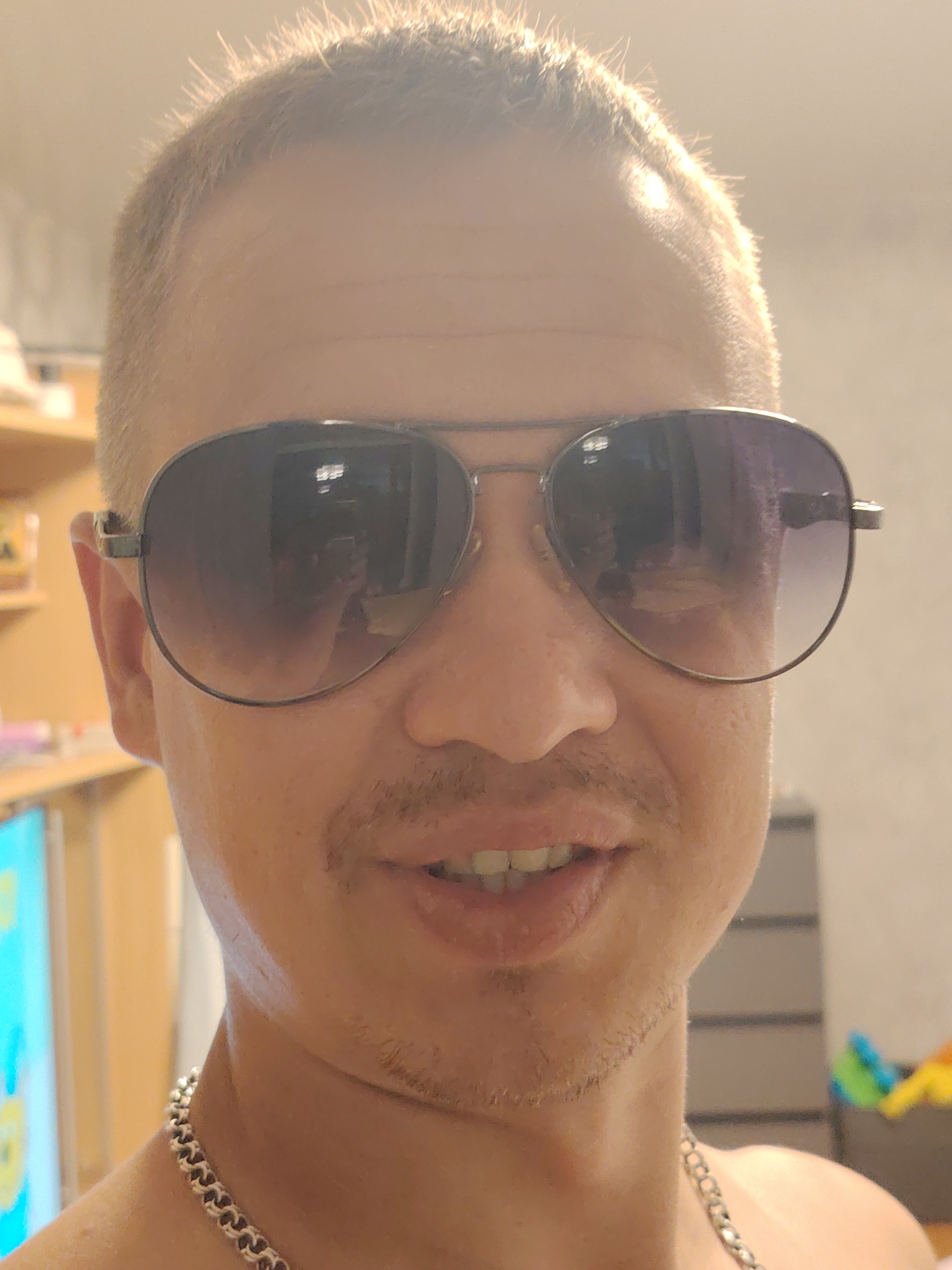 A man with short, blond hair and a mustache is wearing aviator sunglasses. He has a silver chain around his neck. The background appears to be an indoor setting with shelves and some blurred objects. The lighting is warm and seems to be coming from the right side of the image. The man is looking at the camera and his mouth is slightly open, revealing his teeth. The image is a close-up, focusing on the man's face and upper chest. No specific location in Chaikovsky, Russia can be identified.