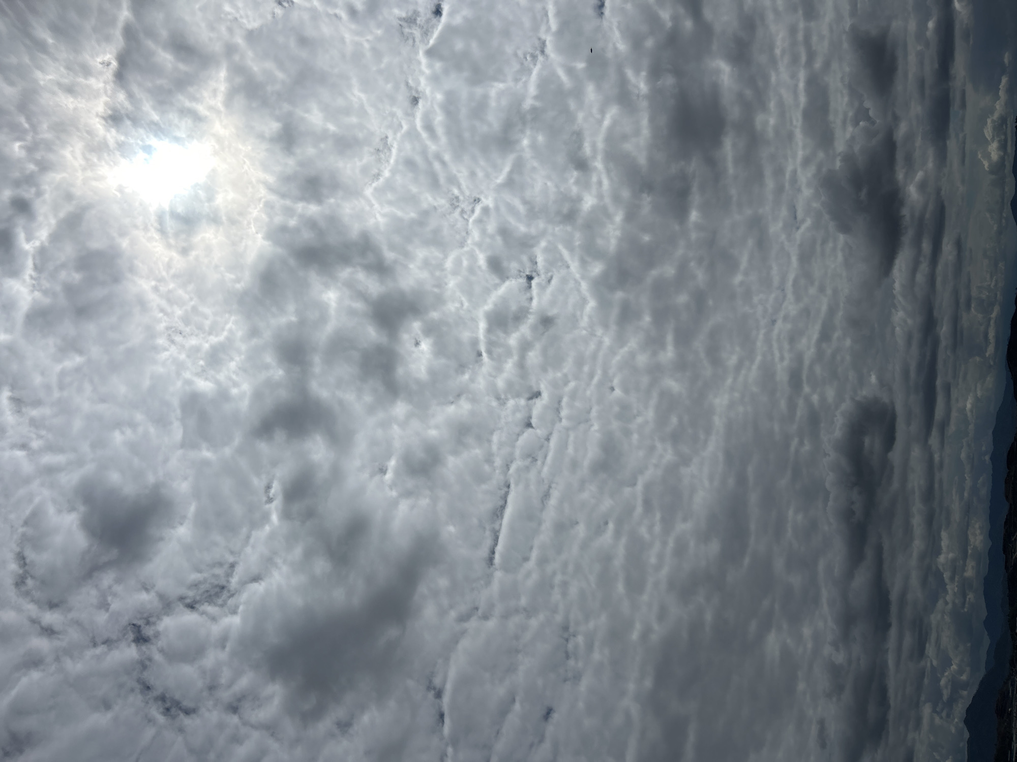 The image depicts a cloudy sky with the sun partially obscured by clouds, located above the city of Port Moresby, Papua New Guinea. The clouds are varied in density, with some appearing thick and dark, while others are lighter and more scattered, allowing a bright portion of the sun to shine through. In the distance, a silhouette of hills or mountains can be seen against the lower horizon, suggesting a natural landscape. The lower part of the image shows a faint outline of what might be urban development or structures on the land. There are no visible people, specific objects, or actions taking place within the frame; the focus is entirely on the atmospheric conditions and the expansive sky. The lighting suggests it could be daytime, possibly late morning or early afternoon, with the sun's intensity filtered by the cloud cover.