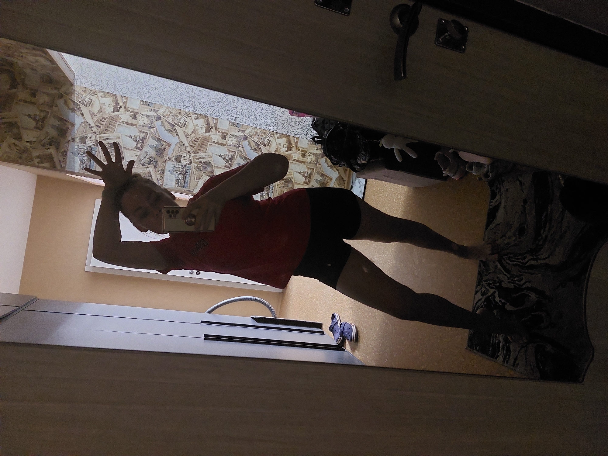 A person is reflected in a mirror. The person, positioned in the reflection, is wearing a red shirt and black shorts and holding up their hand. The background includes wallpaper featuring depictions of landmarks. To the right of the reflection, a rug is visible. A pair of blue Crocs is also seen in the reflection. This image appears to have been taken indoors in a residential setting, possibly in an apartment in Chaikovsky, Russia, given the wallpaper and Crocs.