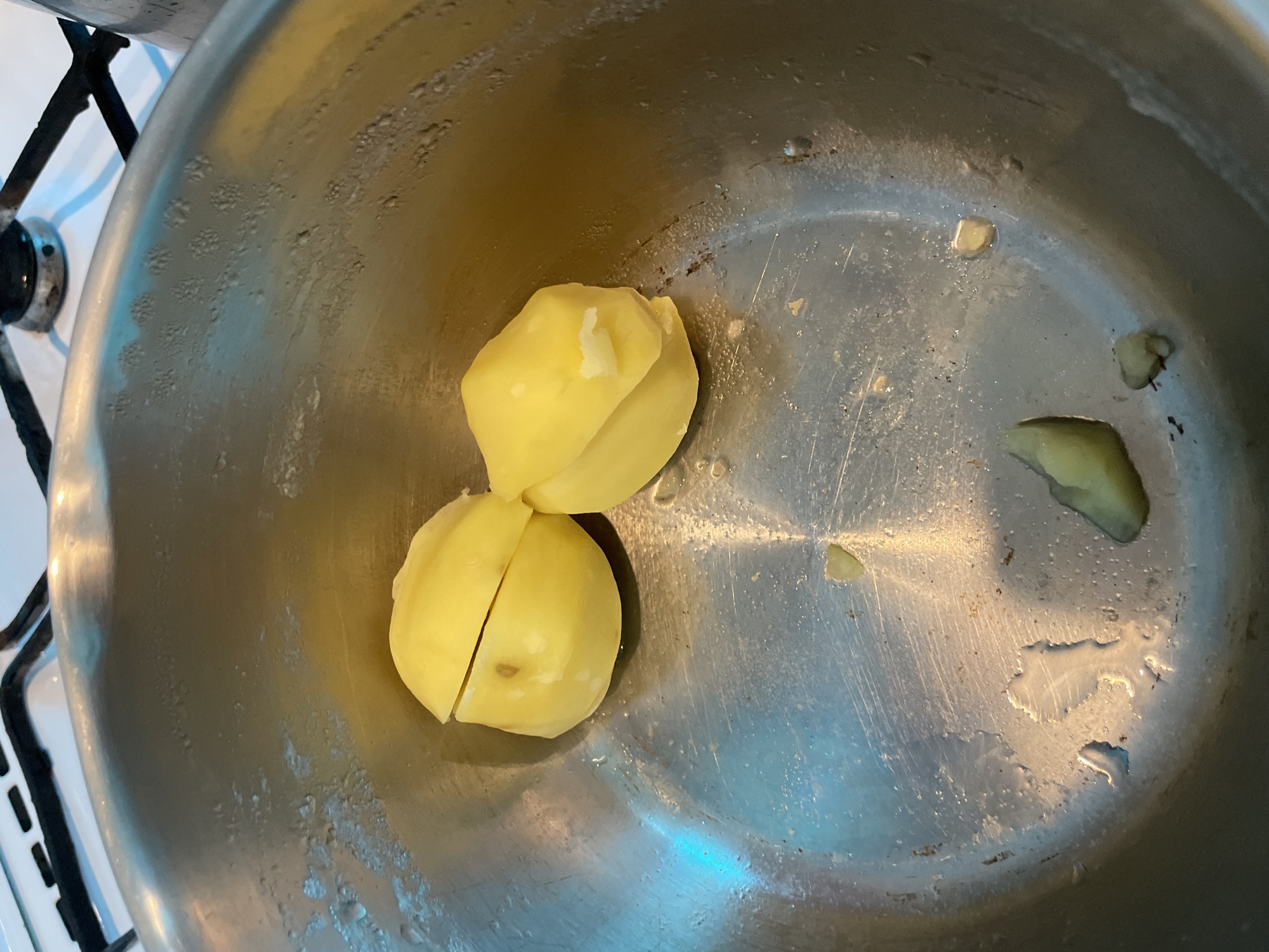 A top-down shot shows a metal pot with two peeled and cut potatoes inside. The pot is on a stove, partially visible in the background. There is some water and small potato pieces scattered around the bottom of the pot. There is no information indicating the location is Olginka, Russia.
