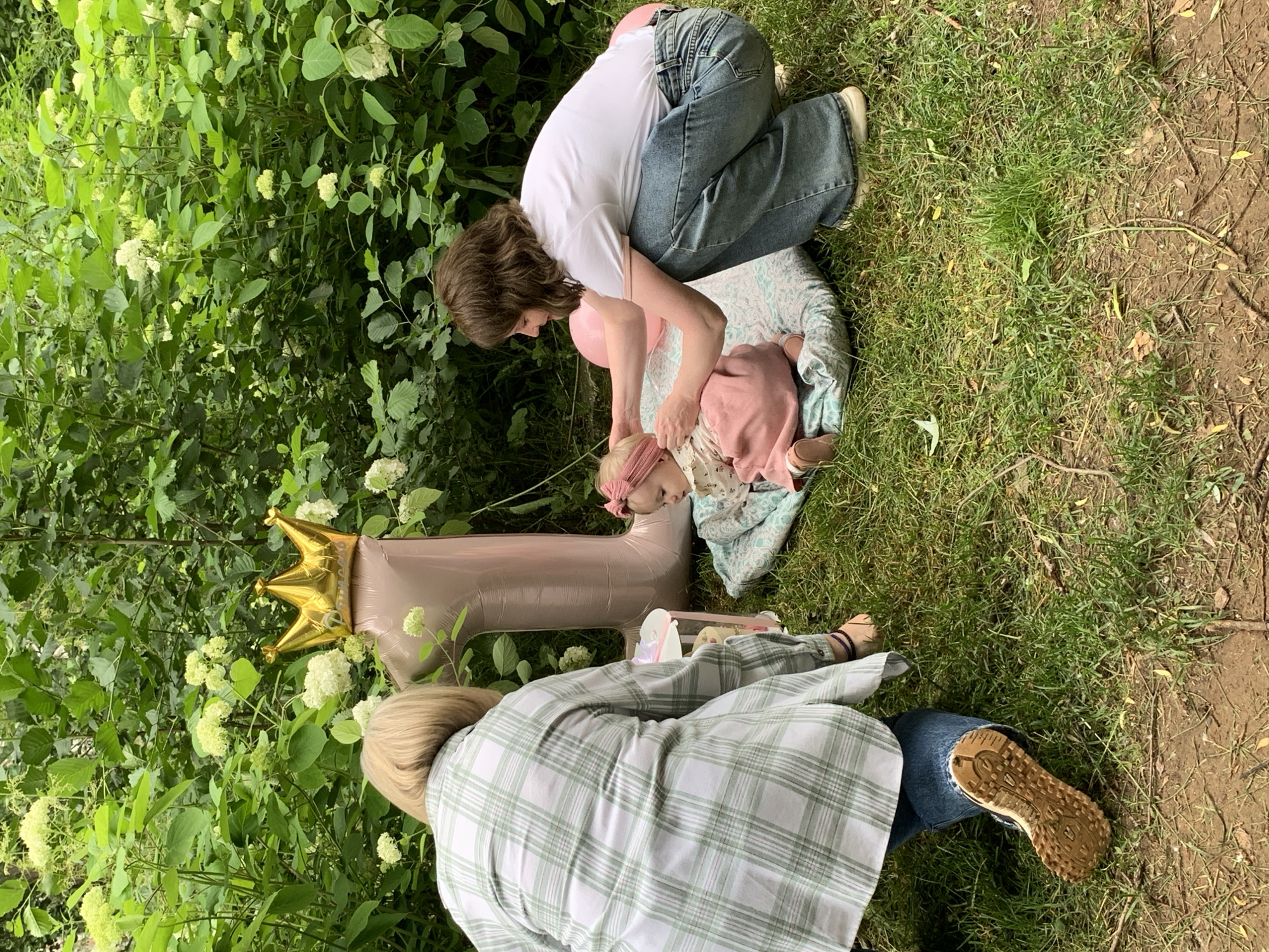 In a grassy outdoor setting, possibly a park or backyard, two women are interacting with a baby. A large pink balloon in the shape of the number '1' topped with a gold crown balloon is positioned behind the baby. One woman, wearing a green and white plaid shirt and blue jeans, is kneeling and appears to be tending to something on a small table. The second woman, in a white t-shirt and blue jeans, is crouched and adjusting the baby's headband. The baby is wearing a pink outfit and is seated on a patterned blanket. The background is filled with lush green foliage and white flowers, suggesting a spring or summer day. The lighting indicates it is daytime. There is no specific indication of the city of Chaikovsky, Russia, in the image.