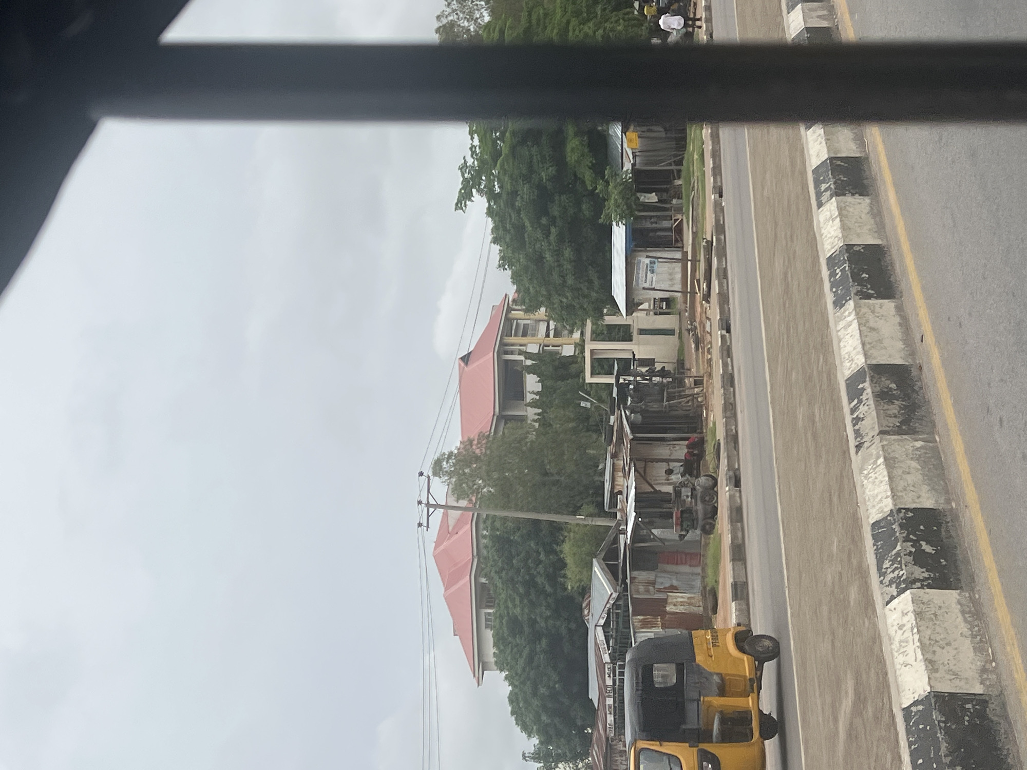 The image depicts a street scene in Jalingo, Nigeria. In the background, a large building with a red roof and cream-colored walls stands amidst lush green trees. In the middle ground, several rustic structures with corrugated metal roofs are visible, some appearing to be small shops or stalls. A yellow three-wheeled vehicle, commonly known as an auto-rickshaw, is parked on the left side of the road. A small generator or engine is positioned in front of one of the stalls. A few individuals are present, one sitting near the generator and another walking in the distance on the right. The foreground shows a road with a distinctive black and white checkered curb. The sky is overcast, suggesting a cloudy day.