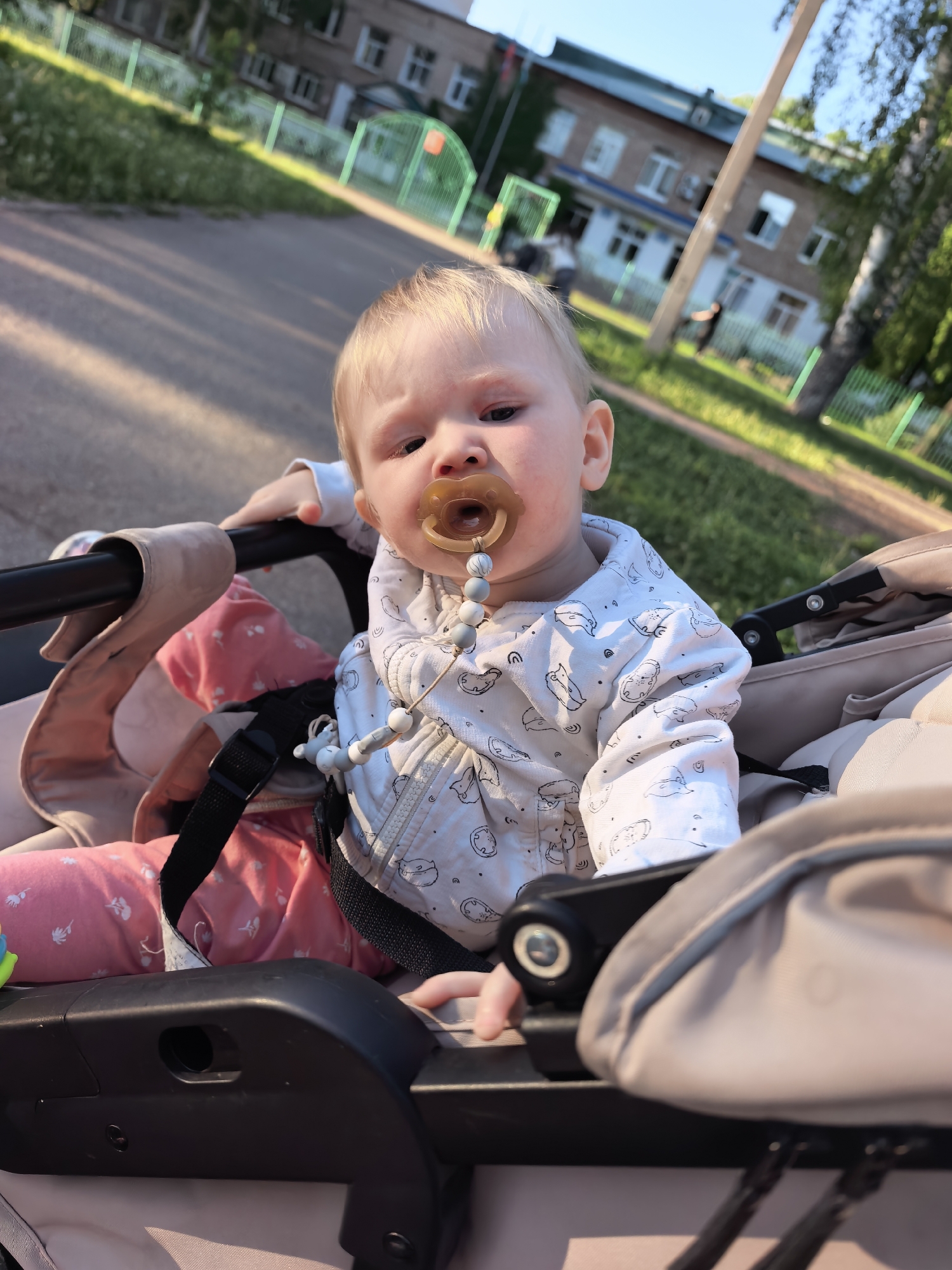 A fair-skinned baby with blonde hair and a pacifier is sitting in a stroller. The baby is wearing a white jacket with a pattern of small figures and pink pants. The stroller is beige with black straps and handle. The location appears to be in Sterlitamak, Russia, as suggested by the presence of a building that is reminiscent of the architecture often found in Russian cities. The background shows a road, a building with several windows, and some trees. The scene is lit by sunlight.