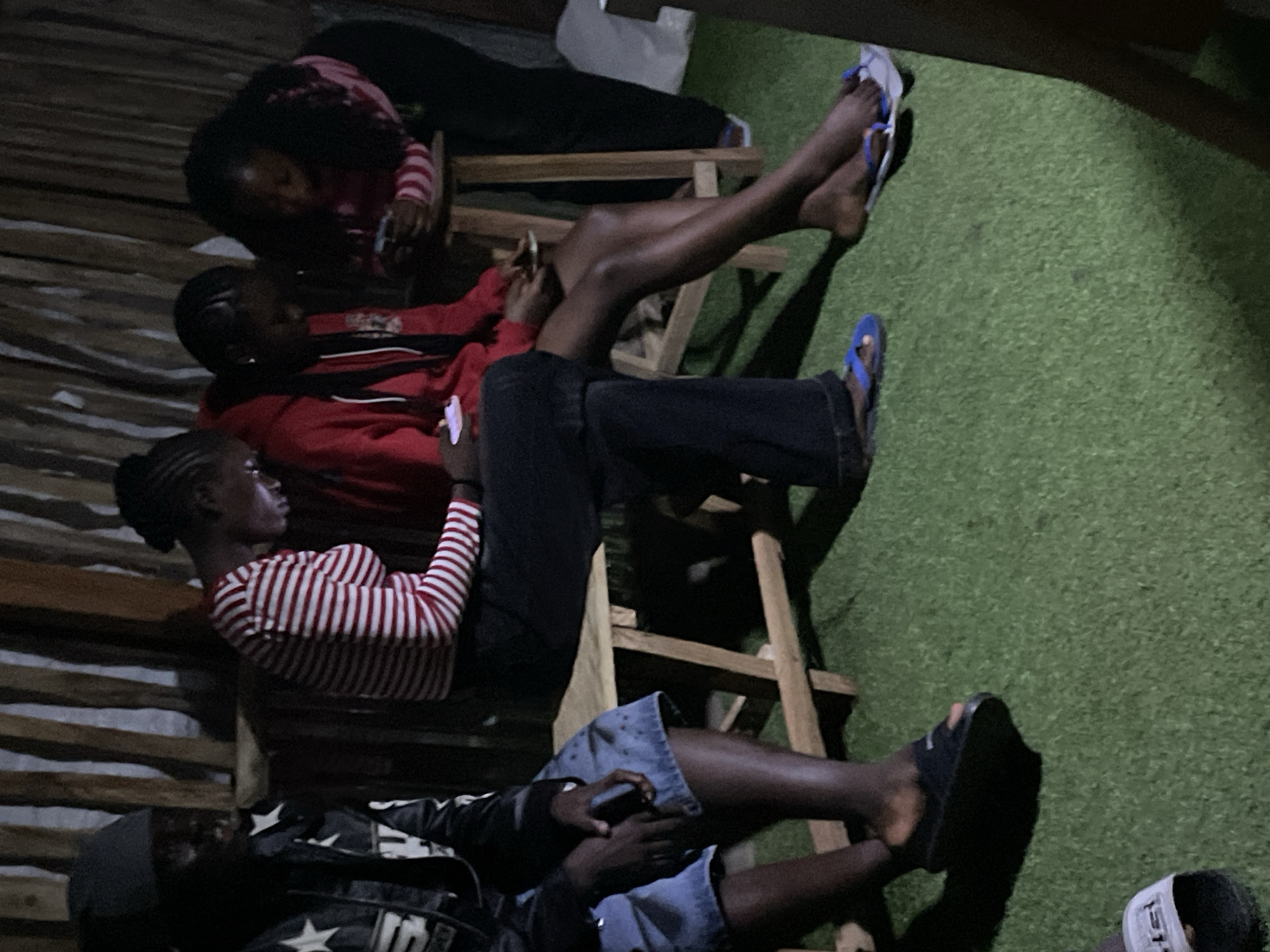 The image captures an outdoor gathering of four young adults, three females and one male, seated on a wooden bench and a stool. The scene appears to be in Durumi, Nigeria, with a backdrop of vertical wooden planks. The individuals are engrossed in their mobile phones, suggesting a shared activity of digital engagement.
The male on the far left is wearing a black jacket with white stars and a dark cap. Next to him, a female in a red and white striped long-sleeved shirt is focused on her phone. Two other females are seated to her right; one in a red hoodie and the other in a pink and white striped top. They too are looking at their phones.
The ground is covered with artificial grass. The lighting in the scene is dim, creating shadows and a slightly murky atmosphere. The composition of the image focuses on the group's interaction with their devices, highlighting a common modern-day social behavior. The presence of sandals on some of the individuals' feet indicates a casual setting. The overall impression is that of a relaxed, informal gathering where technology plays a central role in the participants' attention.