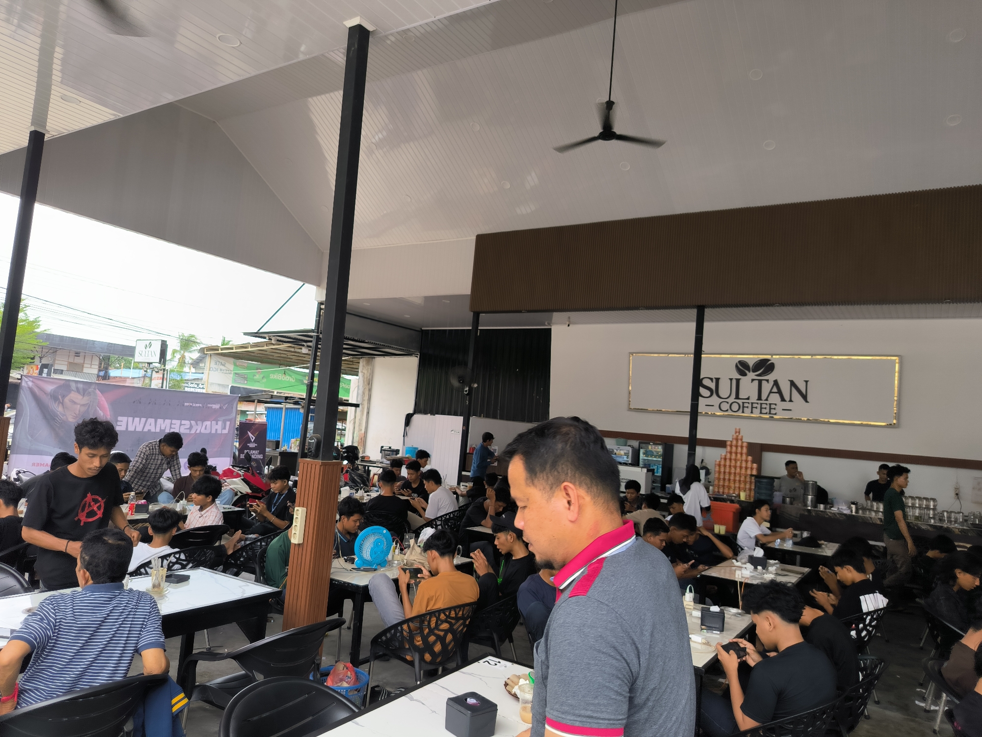 The image depicts a crowded coffee shop, identified by the "SULTAN COFFEE" sign, presumably located in Lhokseumawe, Indonesia. Numerous individuals, mostly young men, are seated at tables, many engrossed in their mobile phones, suggesting gaming or social media engagement. Several individuals are serving and interacting with the patrons. The coffee shop appears to have both covered and open-air seating areas. A banner is visible in the background, possibly advertising an event or game related to the customers' mobile activities. The overall atmosphere seems lively and social.