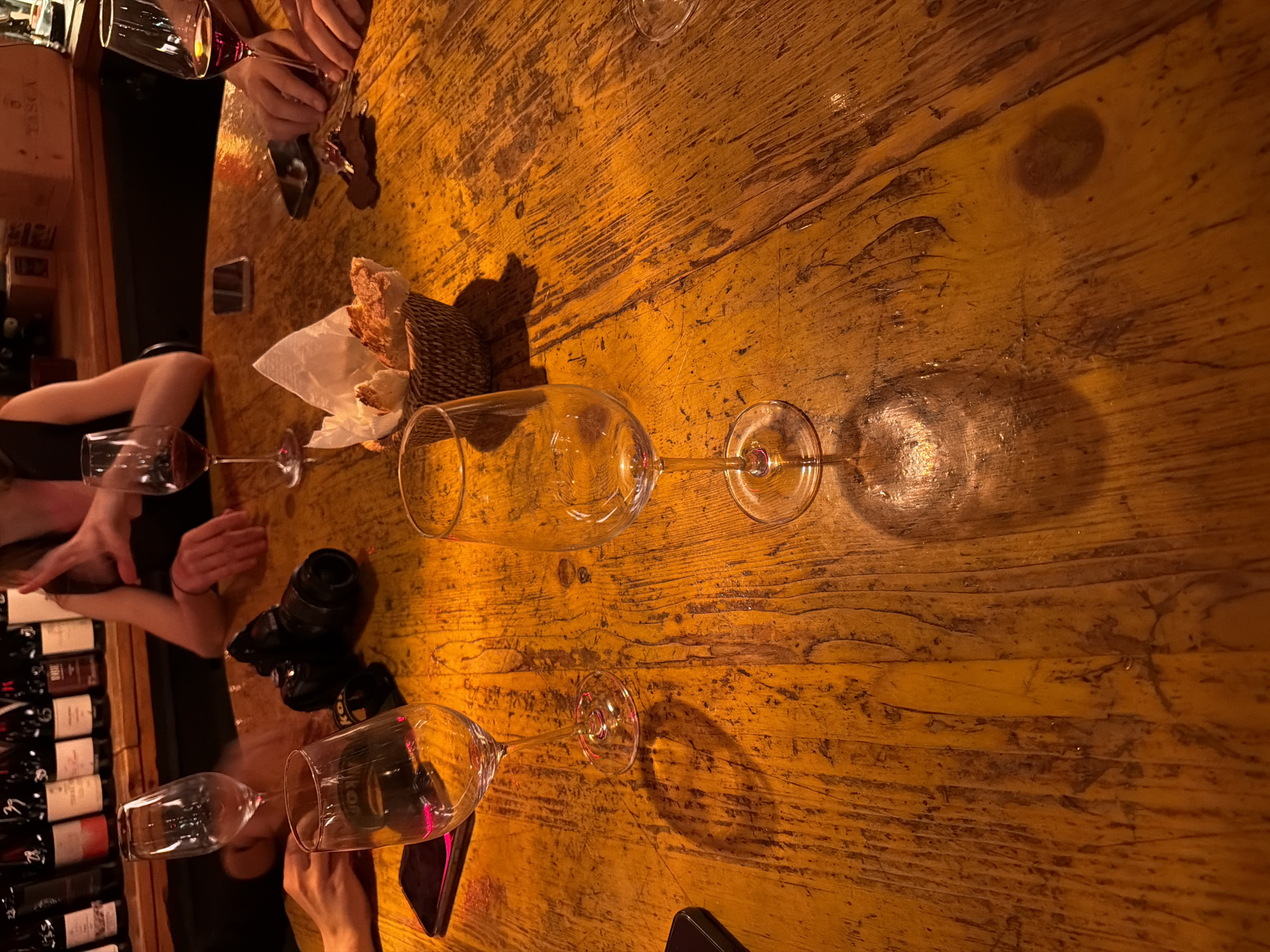 An overhead, low-angle shot shows a collection of wine glasses, a camera, bread, and two cell phones on a rustic wooden table. The table has a warm, amber hue due to the lighting and the wood's natural color. In the background, a rack of wine bottles is visible, suggesting a wine bar or restaurant setting. The scene is illuminated by warm, artificial light that casts distinct shadows across the table.
The image appears to have been taken at a bar or restaurant in Milan, Italy, specifically at a location with a warm, inviting atmosphere.