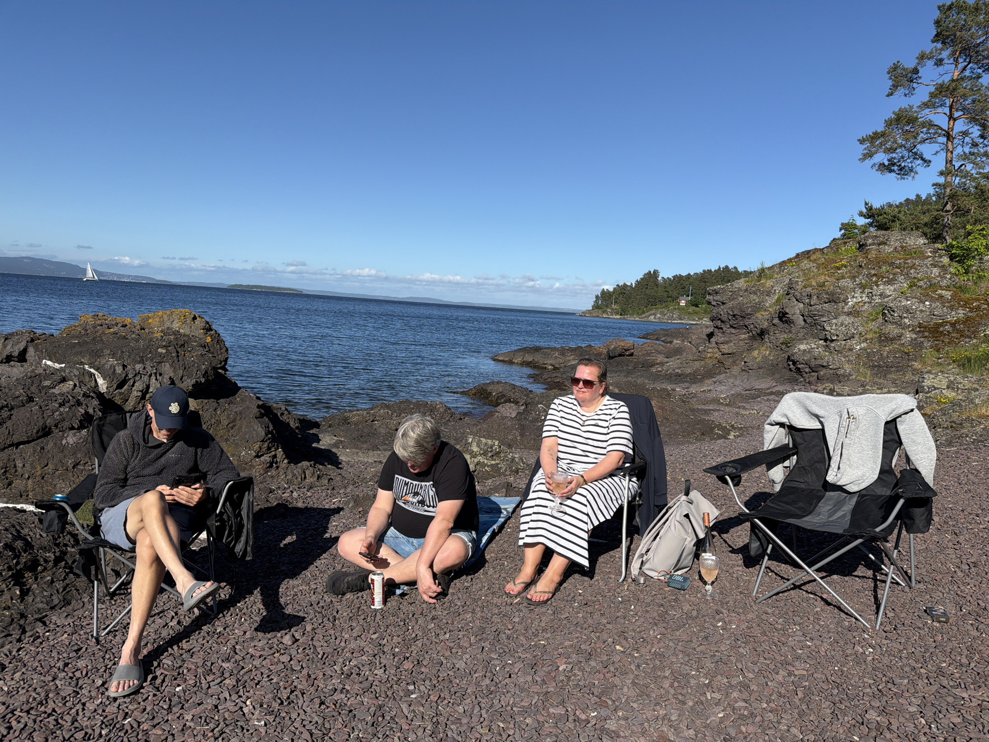 This image depicts a sunny outdoor scene on a rocky and pebbly shoreline in Horten, Norway. Three individuals are present, relaxing near a body of calm blue water, likely a fjord.
On the left, a man wearing a dark baseball cap, a dark textured hoodie, blue shorts, and grey slides is seated in a black camping chair. He is looking down intently at a smartphone held in both hands, with his legs crossed at the ankles. A dark backpack is partially visible behind his chair.
In the center, a person with short, light-colored hair, wearing a black t-shirt with a graphic design and denim shorts, sits cross-legged directly on a blue mat or blanket on the dark pebbles. They are also looking down at a smartphone held in their hands. A white and red beverage can rests on the pebbles to their right.
To the right, a woman wearing large dark sunglasses, a white and black striped maxi dress, and dark flip-flops is seated in a black camping chair. She holds a stemmed glass containing a rosé-colored beverage and is looking towards the left.
An empty black camping chair is positioned to the right of the woman. A grey hooded sweatshirt is draped over its back. On the pebbles beside this empty chair, a grey backpack, a blue and grey JBL portable speaker, and another stemmed glass with a rosé-colored drink are visible. A small, dark rectangular object lies on the ground near the empty chair.
The ground is covered with dark, reddish-brown pebbles. Large dark rocks and boulders line the immediate shore. The background features a wide expanse of deep blue water under a clear, bright blue sky. A white sailboat is visible on the water in the mid-distance to the left, with distant landmasses and mountains faintly visible on the horizon. To the far right, a rocky, vegetated bluff with numerous tall pine trees extends along the coastline. A small building with a red roof is discernible among the trees on the distant right shore. The sunlight is strong, creating distinct shadows on the pebbled ground.