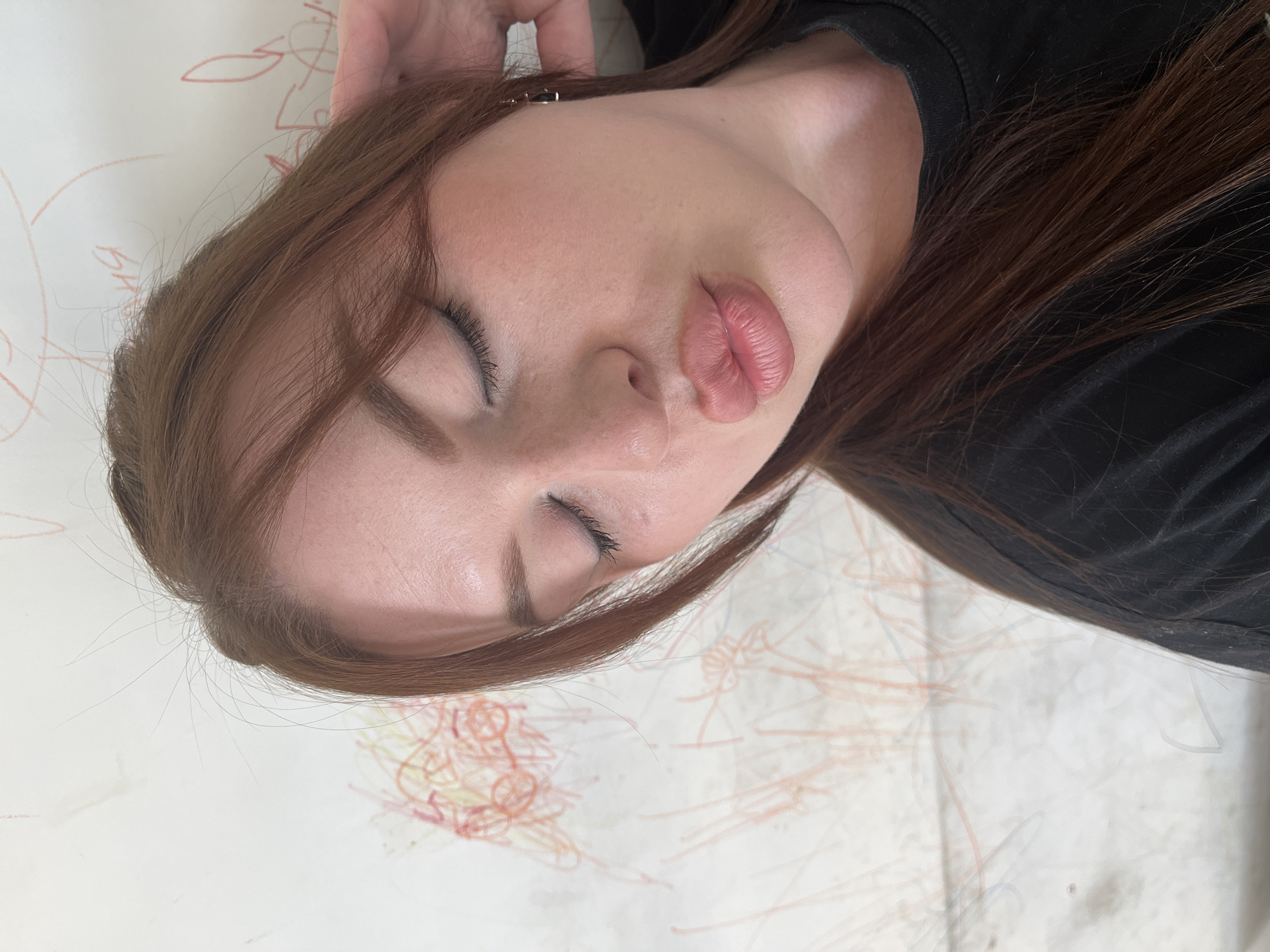 An image, rotated 90 degrees counter-clockwise, depicts a young woman in what appears to be a close-up, head-and-shoulders shot. She has long, dark brown hair that cascades around her head and shoulder. Her eyes are closed, and her lips are noticeably pursed into a "kissing" or "pouting" expression. Her skin is fair, and her eyelashes appear long and dark. She is wearing a simple black t-shirt or top, visible around her neck and shoulder.
The woman is positioned against a light-colored background, likely a wall or a large sheet of paper, which is covered with numerous abstract scribbles and child-like drawings in various shades of red, orange, and faint blue or grey crayon/marker. Some faint, unreadable Cyrillic-like characters can be vaguely discerned amidst the red lines in the upper left portion of the background. A small, dark hair accessory is partially visible near her right ear, obscured by her hair. The lighting is soft and even, highlighting her facial features. The overall mood conveyed is one of playful affection or a moment of whimsical rest. The scene is set in what is identified as Павловка, Russia.