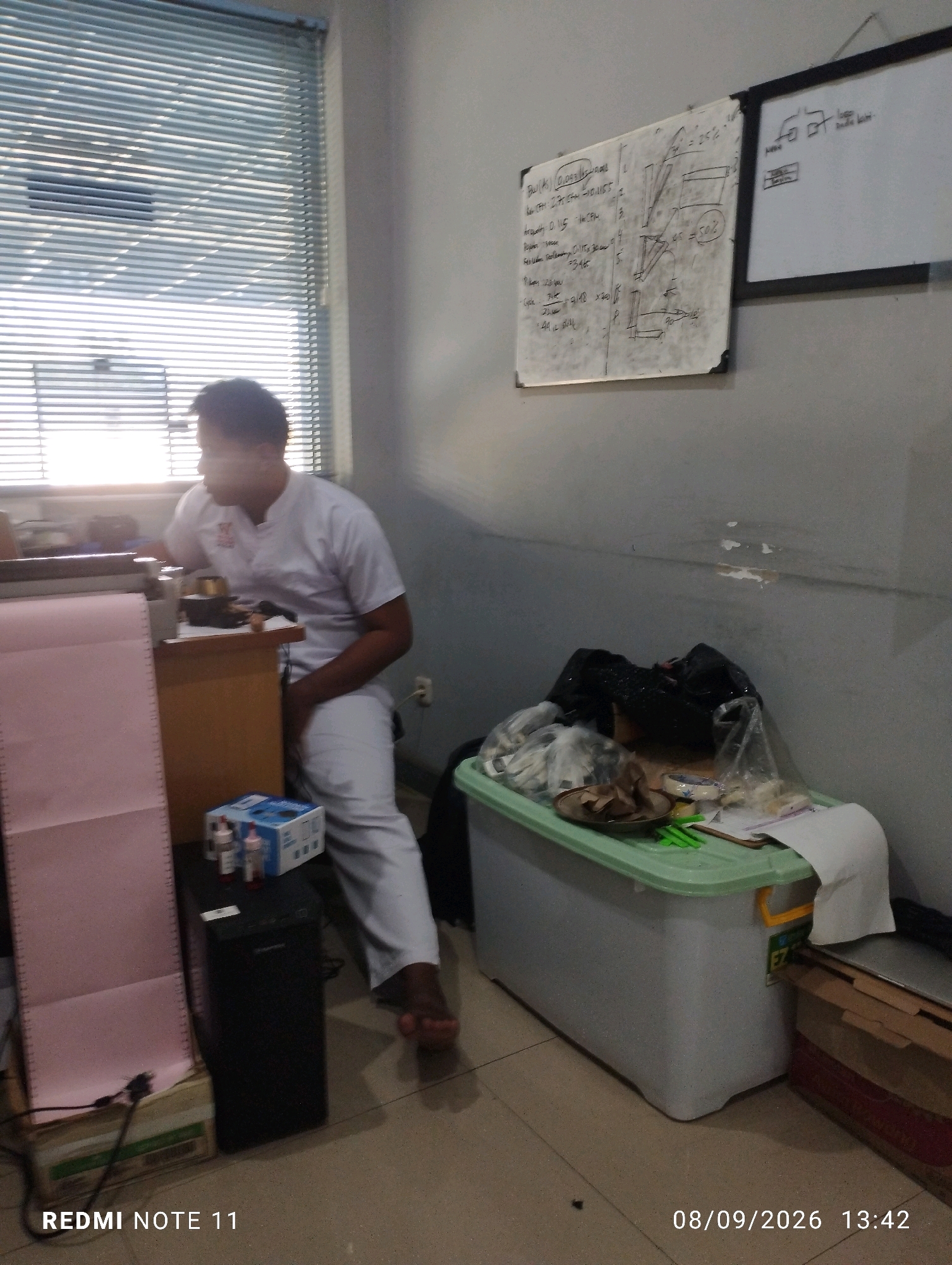 A man wearing a white uniform sits in an office at a desk. To his left, a pink filing cabinet stands against a window covered by blinds. On the desk, a black computer tower is visible, along with a box of medical supplies. To the right of the desk, a large gray and green plastic container is filled with various items, including plastic bags and papers. A whiteboard with diagrams and text hangs on the wall behind the container. The timestamp "08/09/2026 13:42" is visible in the lower right corner. The image captures a typical office environment, with no specific details indicating a location within the city of Bero, Indonesia.