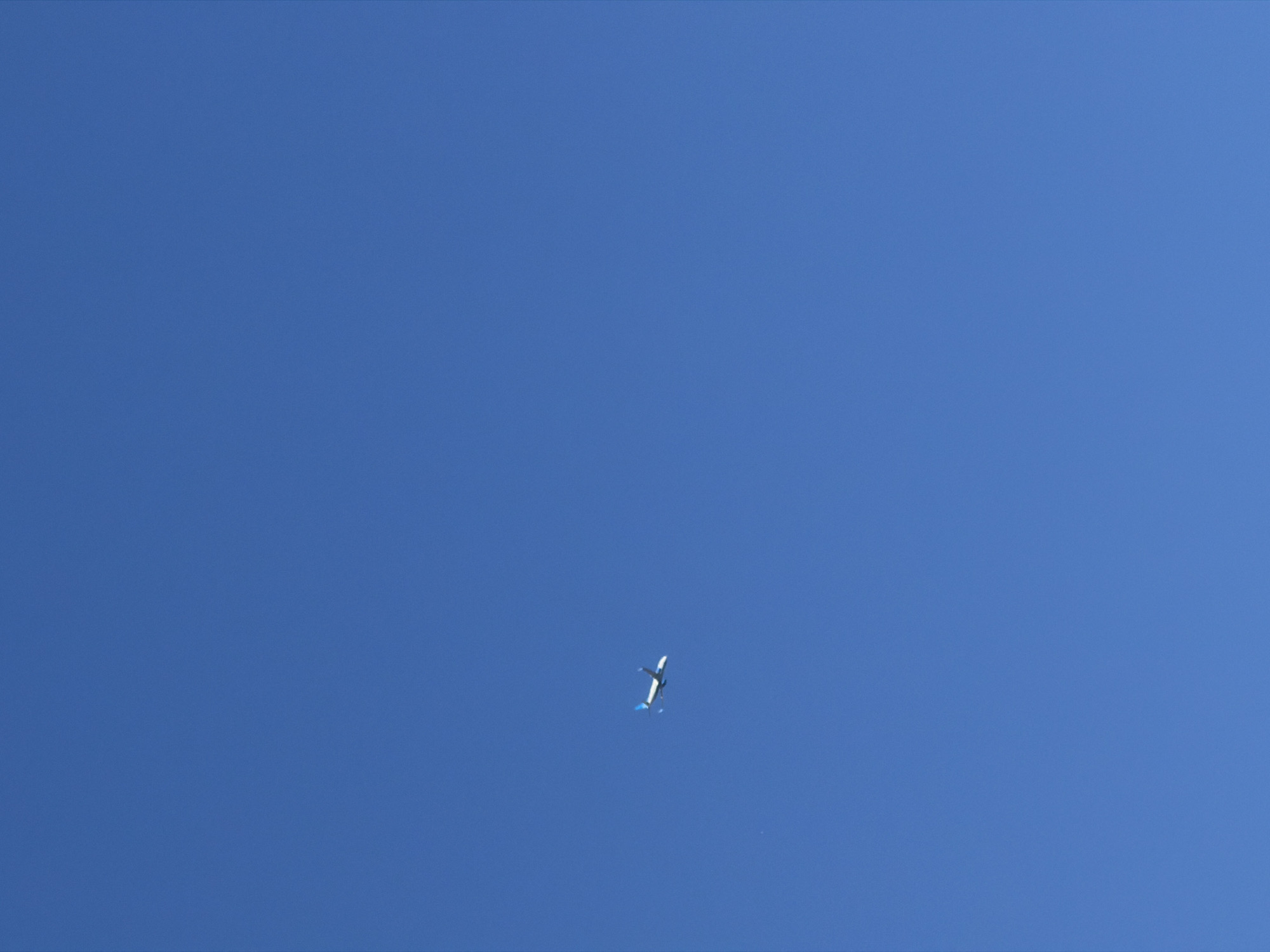 The scene presents a clear, uniform blue sky above the city of Saint Petersburg, Russia. A white passenger airplane, featuring blue accents on its tailfin and possibly wingtips, traverses the upper-left portion of the frame, moving horizontally from left to right. The aircraft is distant and occupies a small fraction of the expansive, unblemished sky. No other subjects, objects, or environmental details are discernible.