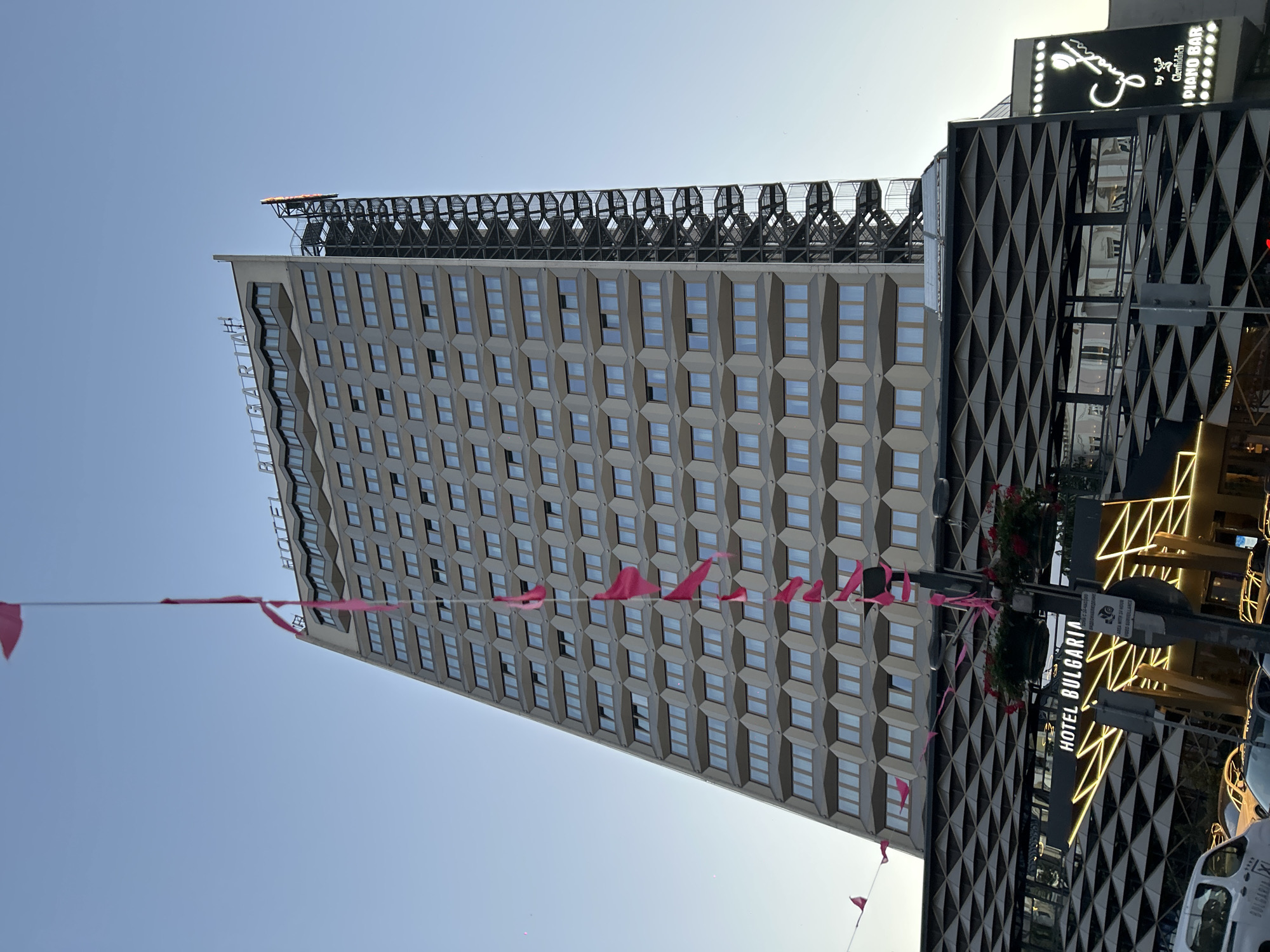 The image shows the Hotel Bulgaria in Burgas, Bulgaria. The hotel is a tall, modern building with a distinctive angled facade. Pink flags are strung across the foreground, fluttering in the breeze. The hotel's name is visible in large letters at the top of the building and also on a lit sign at the base. Below the main hotel building is a modern structure with a geometric pattern facade. To the right of the hotel, a sign for "Sinatra by Glenfiddich Piano Bar" is visible. The sky is clear and blue, suggesting a pleasant day. A fire escape is visible on the side of the hotel. The bottom of the image shows the street level with cars and what appears to be a white van with "BULGARIA" written on its side.