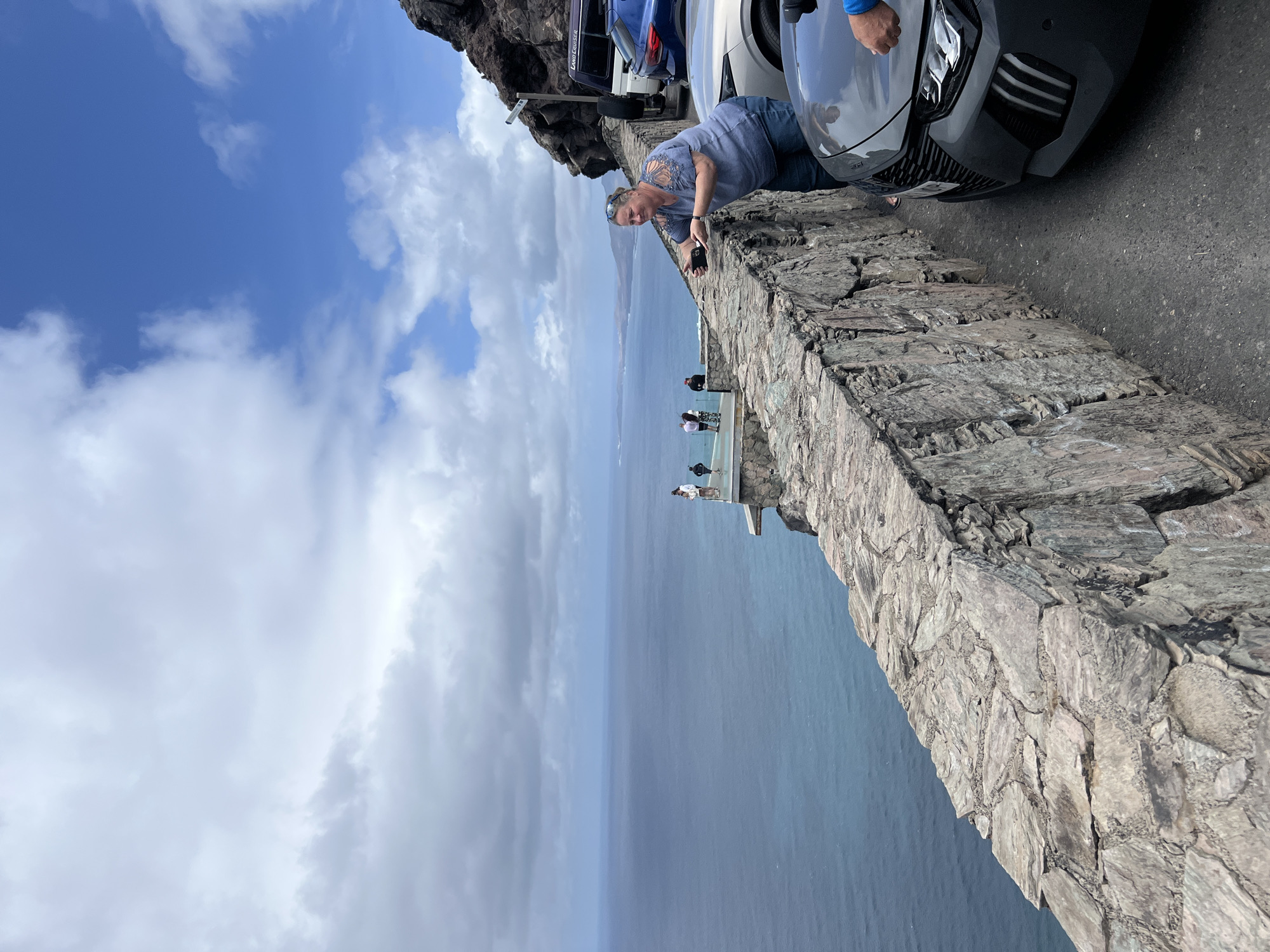 The image captures a scene at the Mirador del Balcón in La Aldea de San Nicolás, Spain. A woman in a blue top and jeans leans over a stone wall, holding a cell phone, possibly taking a photo. Behind her are parked cars, and in the distance, a group of people are visible on a viewing platform. The backdrop consists of the ocean and a partly cloudy sky.