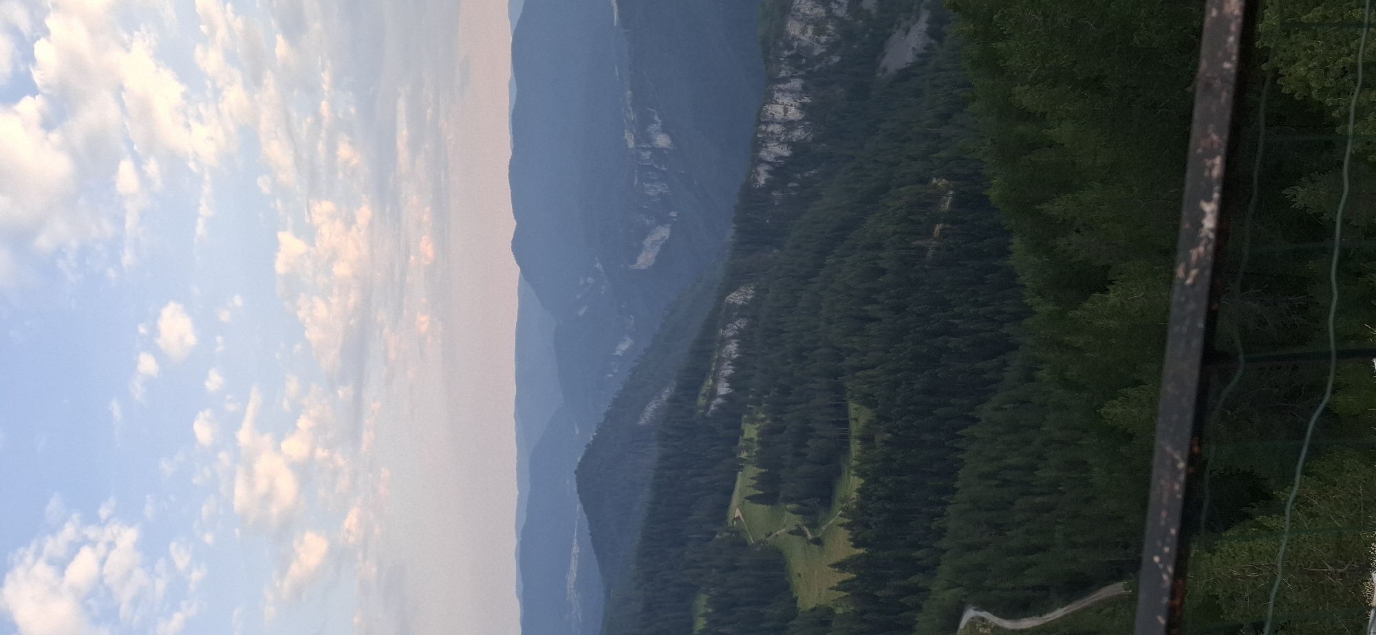 The image displays a panoramic view of a mountainous landscape at dusk. The sky is a soft gradient of blue and pink, with scattered, wispy clouds. Below the sky, layers of mountains recede into the distance, becoming increasingly hazy and blue. The foreground is dominated by a dense, dark green coniferous forest. A winding dirt road is visible in the lower left quadrant, cutting through a small clearing within the forest. The image appears to be taken from a high vantage point, possibly a viewpoint or balcony, as indicated by a metal railing in the bottom part of the frame. The scene depicts a natural, undeveloped environment. There are no people or animals visible in the image. The location is not identifiable as the city of Gyovren, Bulgaria, as no specific landmarks or urban features are present.