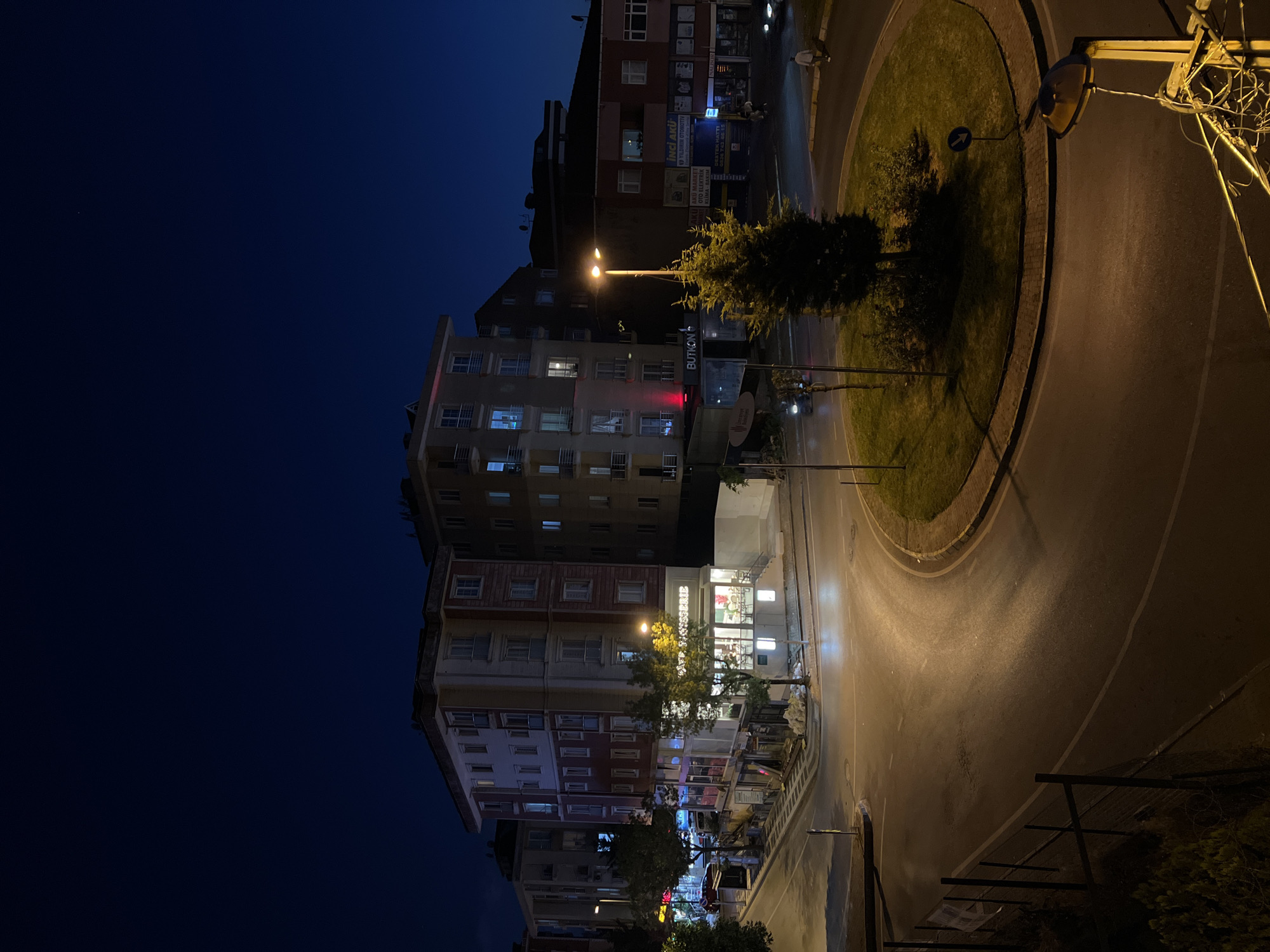 This is an outdoor, nighttime photograph taken from an elevated perspective, likely a building, overlooking a street intersection and a roundabout in Ümraniye, Turkey. The sky is a deep, dark blue, indicating twilight or early night.
In the foreground, a dark metal fence borders a patch of vegetation. The street surface is asphalt, wet and reflective, with tire marks visible. A rounded traffic island is present in the center of the roundabout, covered in grass and featuring a small tree and bushes, illuminated by a streetlamp. Another streetlamp with exposed wiring is visible in the lower right corner.
Several multi-story buildings line the streets. The building directly ahead and to the left has a light-colored facade with rows of windows and balconies. A sign with the text "Cafe Patisserie" is illuminated in white lettering on its ground floor. To the right of this building, another structure is visible with similar architectural features. Further to the right, a red-roofed building has illuminated windows and storefronts on its lower level, displaying signs such as "İNCİ AKÜ" and "AKÜ MARKET."
Vehicles are sparsely present; a motorcycle is seen on the left side of the intersection, and a car is parked near the red-roofed building. A lone pedestrian is walking on the sidewalk on the right side of the image. Construction debris, including white bags, is piled near the base of one of the buildings. The overall scene depicts a quiet urban environment at night.