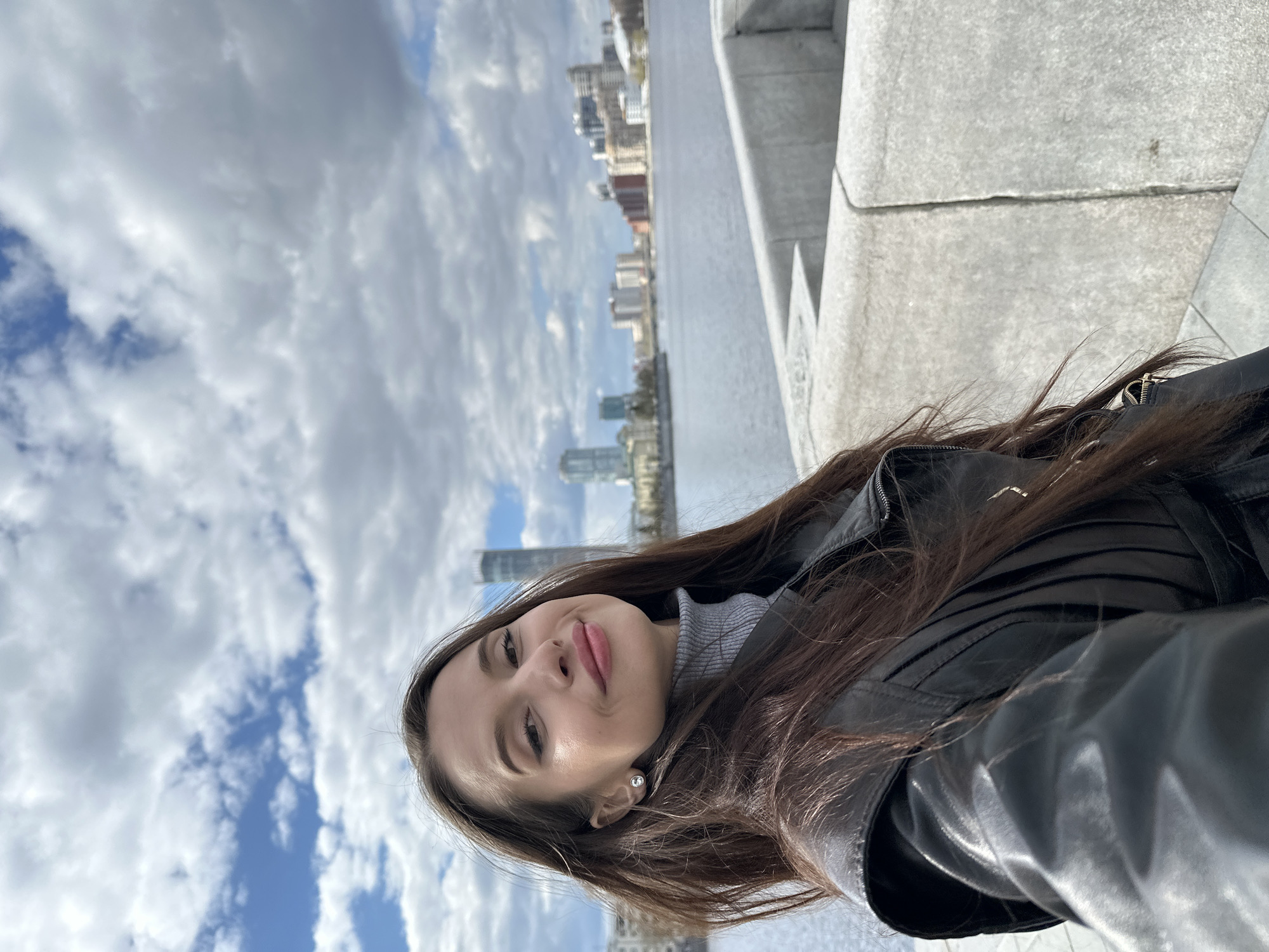 A young woman with long brown hair and fair skin is seen in a selfie. She is wearing a black leather jacket over a gray turtleneck sweater and a small pearl earring. The background consists of a cityscape, a body of water (likely a river or lake), cloudy skies, and a section of concrete architecture.
Based on the architectural style and skyline visible in the background, the location is likely Yekaterinburg, Russia.