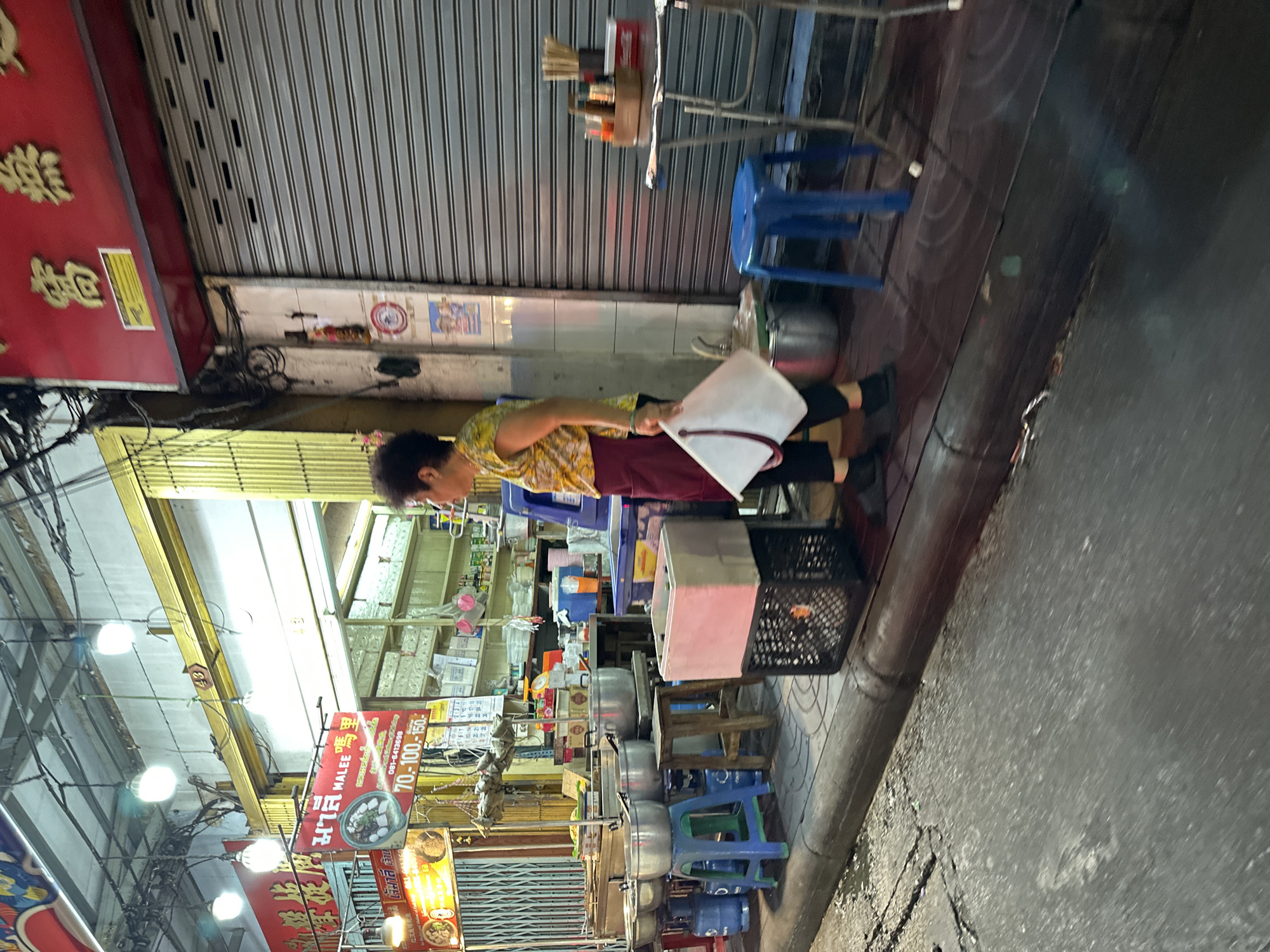 This vertical street-level photograph captures a nighttime scene, likely in Bangkok, Thailand, featuring a woman attending to a street food stall. The woman, positioned centrally, wears a patterned yellow shirt and a maroon apron, and is holding a white piece of paper. She appears to be either setting up or managing the food stall.
The stall setup includes several large metal cooking pots on what appear to be portable stoves or stands, accompanied by blue plastic stools and wooden tables. On one small table, there are various condiments and utensils. In the foreground, part of a black and a pink crate are visible. The ground transitions from a tiled sidewalk to an asphalt road on the right.
The background shows closed corrugated metal shutters of shops, indicating the evening or night-time setting. Bright fluorescent lights illuminate the area, reflecting off the shiny surfaces. Visible text on banners and signs includes Thai and Chinese characters. One prominent sign displays "MALEE 嗎里" (Malee) and a phone number "081-6413698", along with price points "70.-100.-150-", suggesting the cost of dishes offered. Other numbers like "151617" and "43" are also present on different signs. The vibrant signage and utilitarian setup are characteristic of a bustling Asian urban street food environment.