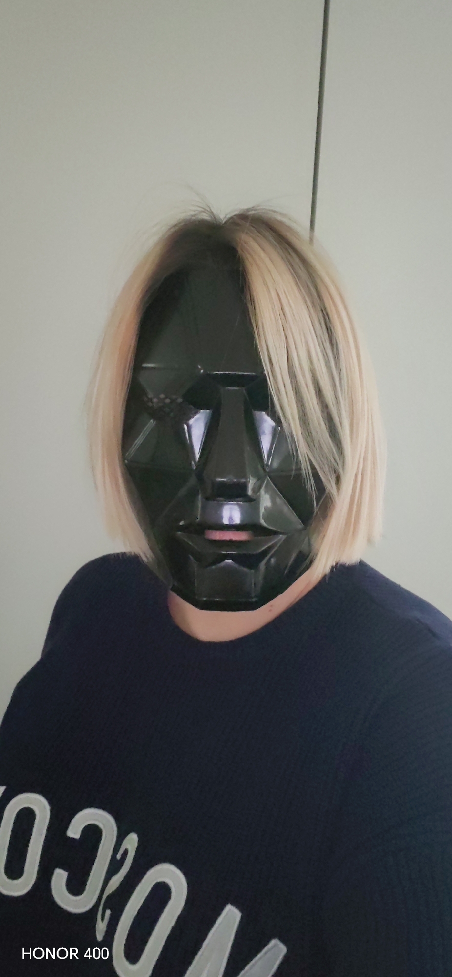 A person with blonde hair is wearing a black geometric mask that covers their face from the forehead to just below the mouth. The person is wearing a dark blue sweater with white lettering partially visible on the lower left of the frame. The background is a plain, light-colored wall. A thin, dark vertical line is visible behind the person's head. The lower left corner of the image contains the text "HONOR 400".