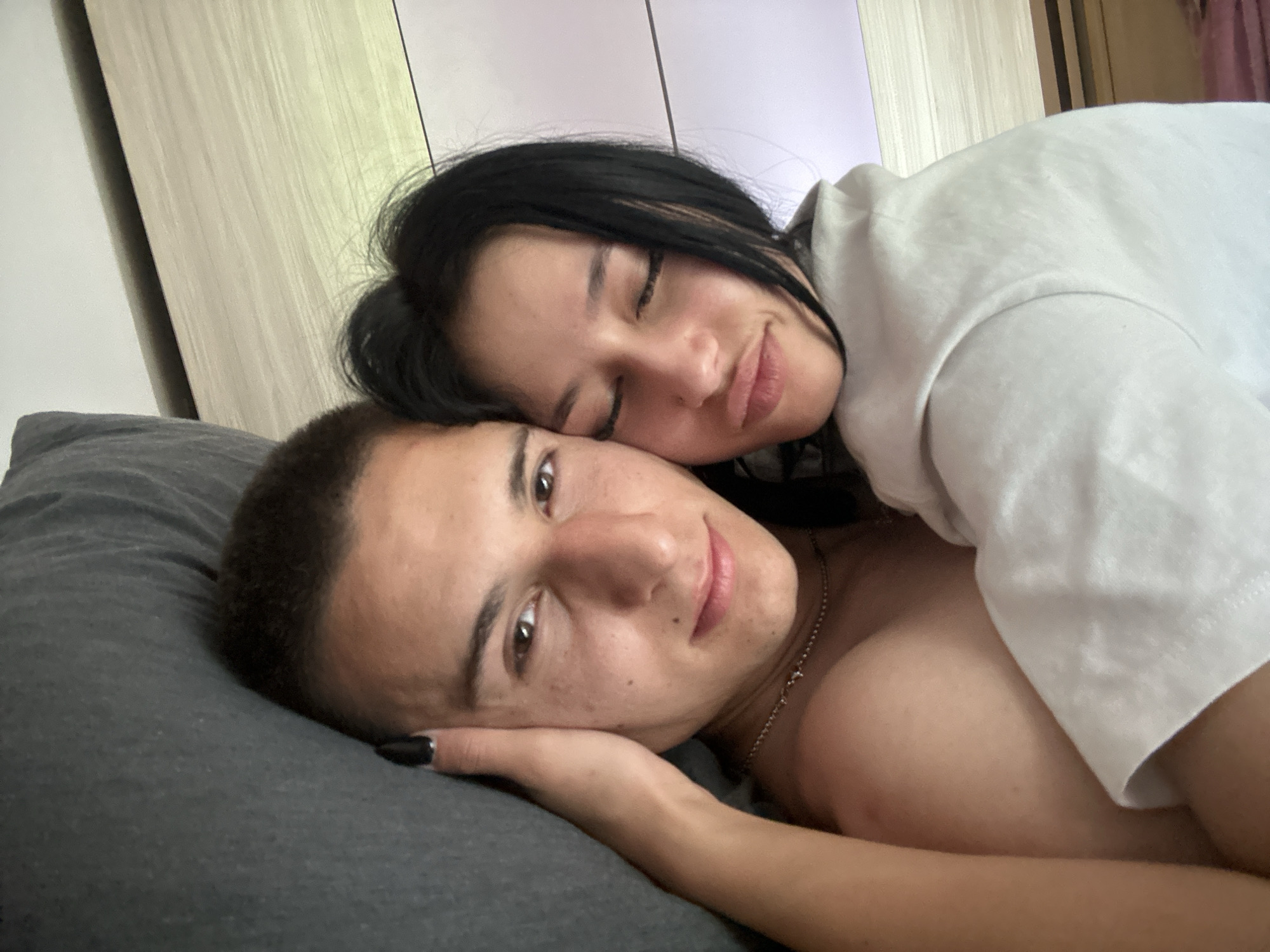 A young adult couple is lying together on a gray pillow. The man has short dark hair and is looking at the camera. The woman has long dark hair and is lying with her head on his shoulder, eyes closed. She is wearing a white t-shirt. The man is shirtless and wearing a thin necklace. A partially visible wardrobe stands in the background. The location is likely indoors. The setting in Burgas, Bulgaria, is not possible to verify from this image.
