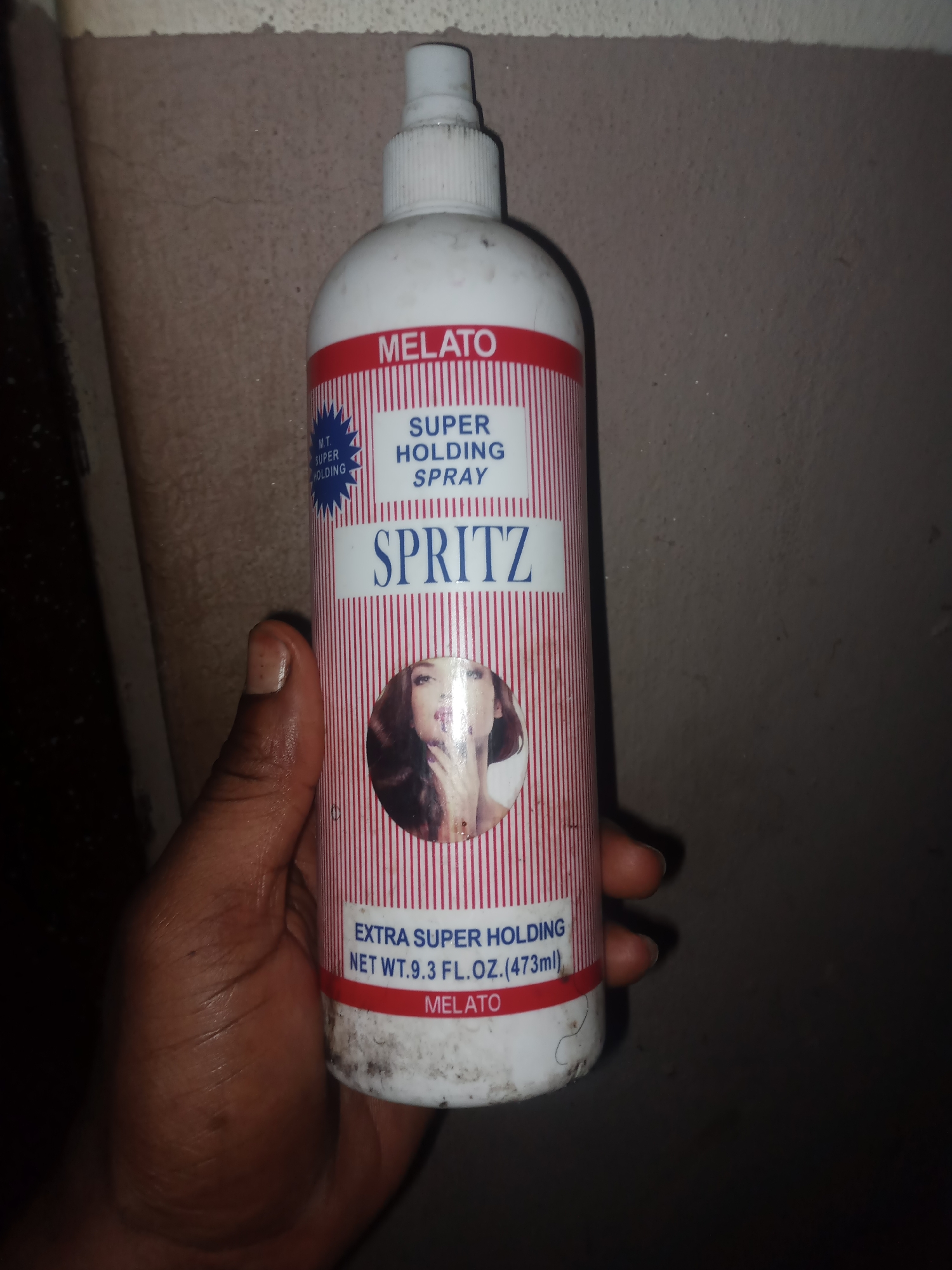 A hand holds a white spray bottle with red and white striped labeling. The label displays "MELATO" in red, followed by "SUPER HOLDING SPRAY" and "SPRITZ" in blue text. A circular image of a woman's face is on the bottle. Below this, "EXTRA SUPER HOLDING NET WT. 9.3 FL.OZ.(473ml)" is printed, along with "MELATO" again. The background is a rough, textured, gray wall. The image appears to be taken indoors, possibly in a storage area, given the texture of the wall and the presence of what looks like dust or grime on the bottle. The lighting is dim, casting shadows and highlighting the texture of the wall and the subject's hand. There is no discernible indication of the specific location within Jauro Yinu, Nigeria.
