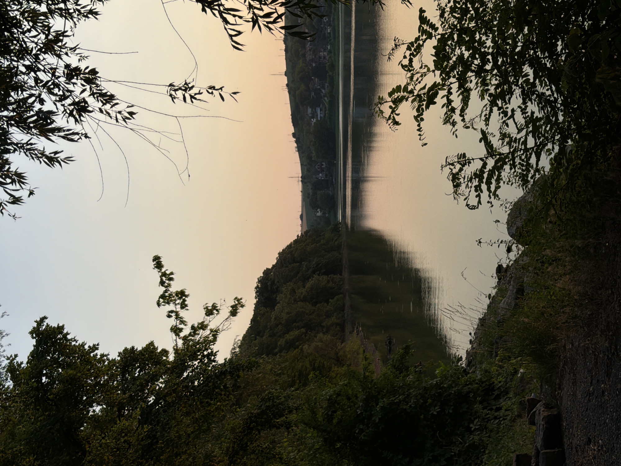 This media file depicts a serene natural landscape at Nikolovo, Bulgaria, likely during dawn or dusk due to the soft, warm lighting. The scene is dominated by a calm body of water, reflecting the sky and the surrounding lush greenery. Silhouetted trees frame the view, with their branches and leaves creating a natural border.
The foreground is characterized by dense foliage, with dark green leaves and branches obscuring parts of the landscape. On the right side, a rocky embankment is visible, covered in patches of grass and small plants. The water's surface is smooth, creating a mirror-like reflection of the tree-lined hill on the opposite bank and the pastel-colored sky.
The hill across the water is covered in dense trees, with a few scattered houses or buildings visible amidst the foliage. The overall impression is one of peace and tranquility. The time of day suggests a quiet moment, possibly early morning or late evening, as indicated by the gentle light and the calmness of the water. The weather appears to be clear, contributing to the peaceful atmosphere. No text is visible in the image.