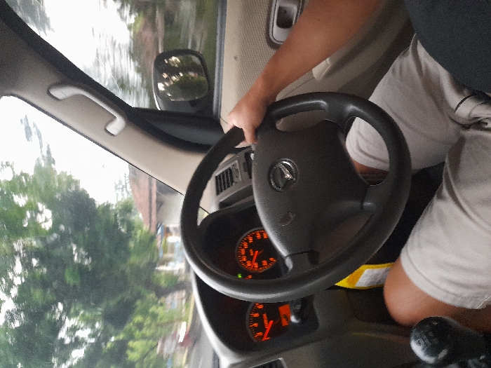 The image depicts the interior perspective from the driver's seat of a moving vehicle in Cangkringrandu, Indonesia. The driver, wearing light-colored shorts and a dark shirt, has their right hand gripping the top right quadrant of a black three-spoke steering wheel featuring a circular emblem, likely a Daihatsu logo. The dashboard is visible, with the instrument cluster displaying a tachometer needle between 1,000 and 2,000 RPM, and a speedometer indicating approximately 40-50 km/h, both with visible red warning lights. A black manual gear shift knob is positioned to the lower left of the frame. A yellow object is situated between the driver's knees and the center console. Through the windshield and the right side mirror, a blurred outdoor environment is visible, comprising green trees, foliage, and residential structures with reddish-brown tiled roofs, consistent with vehicle motion. The passenger-side A-pillar with a grab handle is also present.