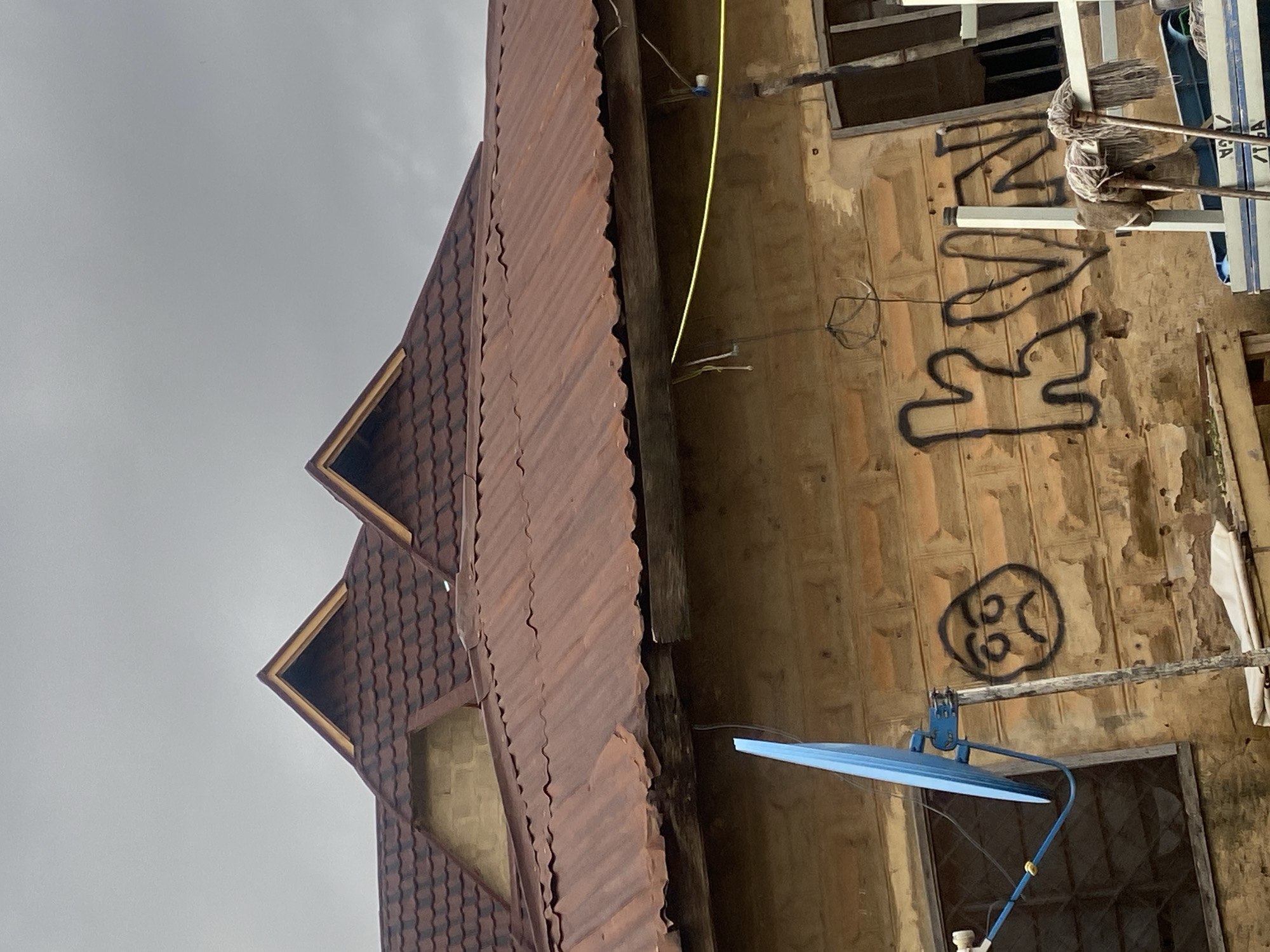 This image shows the side of a building with a corrugated iron roof. The wall of the building is made of light-colored bricks and has graffiti on it, including what appears to be stylized letters and a drawing of a face with a distressed expression. A blue satellite dish is mounted on the wall. There are also some brooms leaning against the wall near the top of the image. The sky is overcast, suggesting it might be a cloudy day. The image is oriented vertically, with the roofline at the left and the wall and satellite dish to the right. The overall impression is one of a somewhat weathered and possibly unfinished or neglected building. There is no clear indication of activity or event taking place in the image.