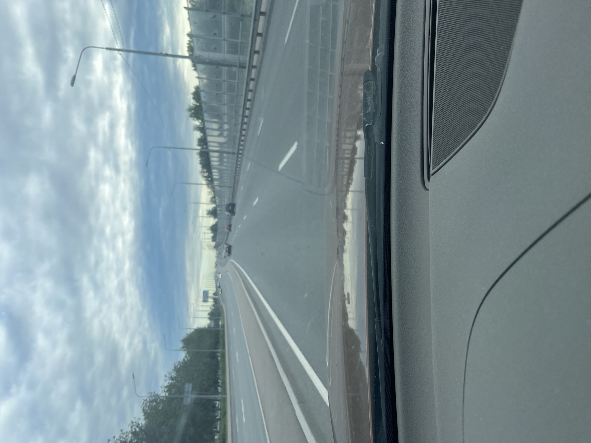 The image is a vertical shot from inside a vehicle looking out at a highway. The scene depicts a road with multiple lanes, separated by white lines. On the left side of the highway, there is a tall sound barrier with a grey, segmented surface, and beyond that, a line of trees and streetlights. The sky is overcast with clouds, suggesting a cool or cloudy day. The time of day appears to be either early morning or late afternoon due to the diffused light.
On the right side of the frame, the interior of the car is visible, showing a dark grey dashboard and a speaker grill. The windshield wipers are visible at the bottom right of the interior. A reflection of the highway and the sky can be seen on the windshield. There are a few cars visible on the highway in the distance, indicating ongoing traffic. The overall atmosphere is neutral, focused on the travel scene and the roadside environment. The location is identified as Saint Petersburg, Russia. No text is visible in the image.