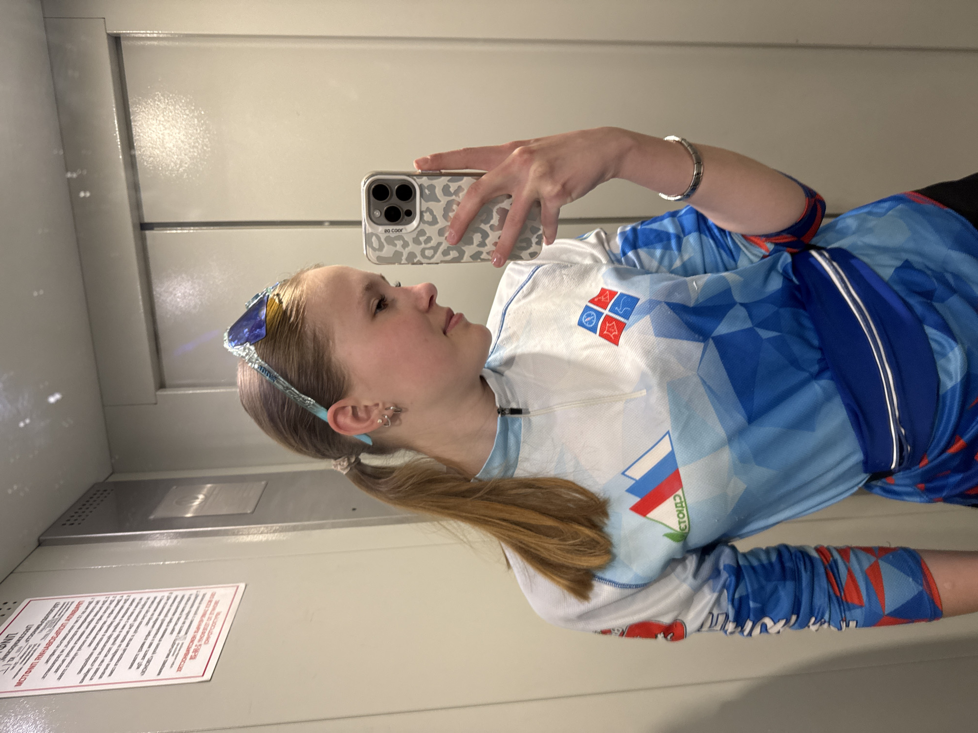 The image shows a person taking a selfie in what appears to be an elevator. The person is wearing athletic gear with a blue and white design featuring what looks like a Russian flag emblem and a logo. The person has a ponytail and a blue headband with glasses on top of the head. The person is holding a phone with a leopard print case. The background suggests an elevator interior, featuring a stainless steel panel and a warning sign in Russian. While there are clues of Russia through the logo, it does not contain information on Chaikovsky, Russia.