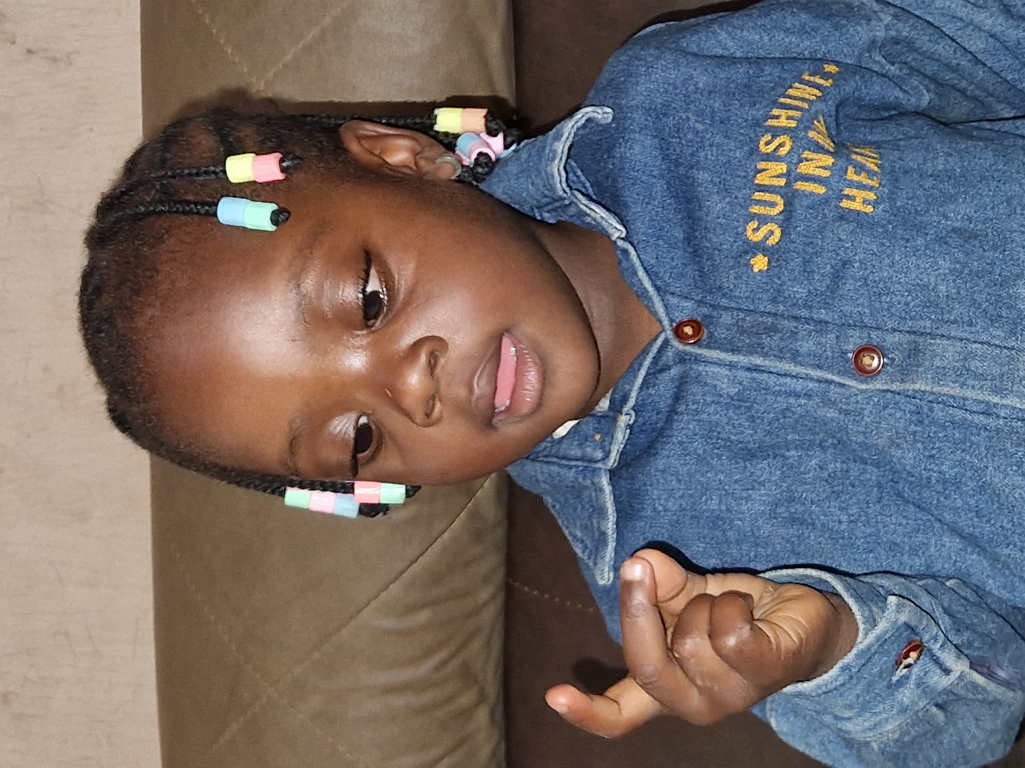 A young Black child with braided hair adorned with colorful beads is shown from the chest up, sitting on a brown couch. The child is wearing a denim jacket with "SUNSHINE IN MY HEART" embroidered in yellow on the left side. The child's left hand is raised, with the index finger extended. The background consists of a textured brown couch and wall. The image appears to have been taken indoors, likely in a residential setting in Jos, Nigeria.