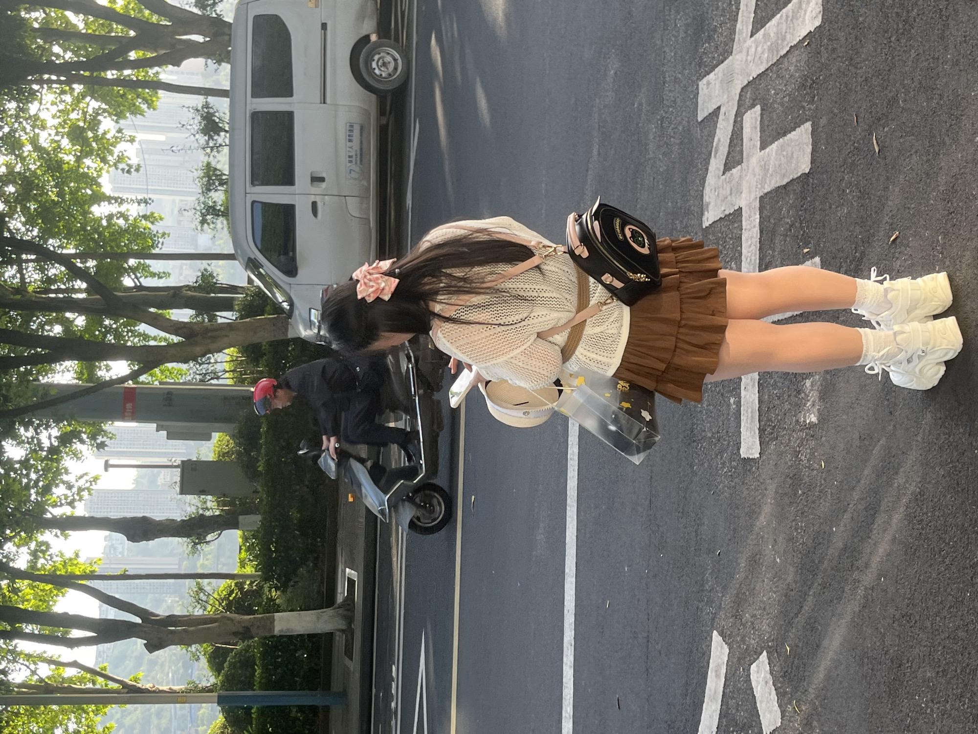 The image depicts a vibrant street scene on a sunny day in an urban area of Jiangjin, China. The primary subject is a young woman, seen from the back and left side, standing on a paved road. She has long, dark hair adorned with a pink bow and is wearing a light-colored, possibly white or cream, open-knit cardigan, a brown pleated mini-skirt, white socks, and white chunky sneakers. She carries a small black handbag with pink accents and a golden chain, and holds a straw hat along with a clear plastic bag containing yellow items.
To the woman's left, a person wearing a dark jacket and a red cap stands next to a dark-colored scooter or small motorcycle. In the background, a white utility van is partially visible. The road features clear white markings, including what appears to be a segment of a large Chinese character, likely "之" (zhī). Green trees line the street, partially obscuring the distant view of numerous tall city buildings, indicating a dense urban or suburban environment. The bright sunlight suggests it is daytime. On the side of the white van, visible text includes "江津" (Jiangjin) and what looks like a partial phone number starting with "17", aligning with the provided location context.
