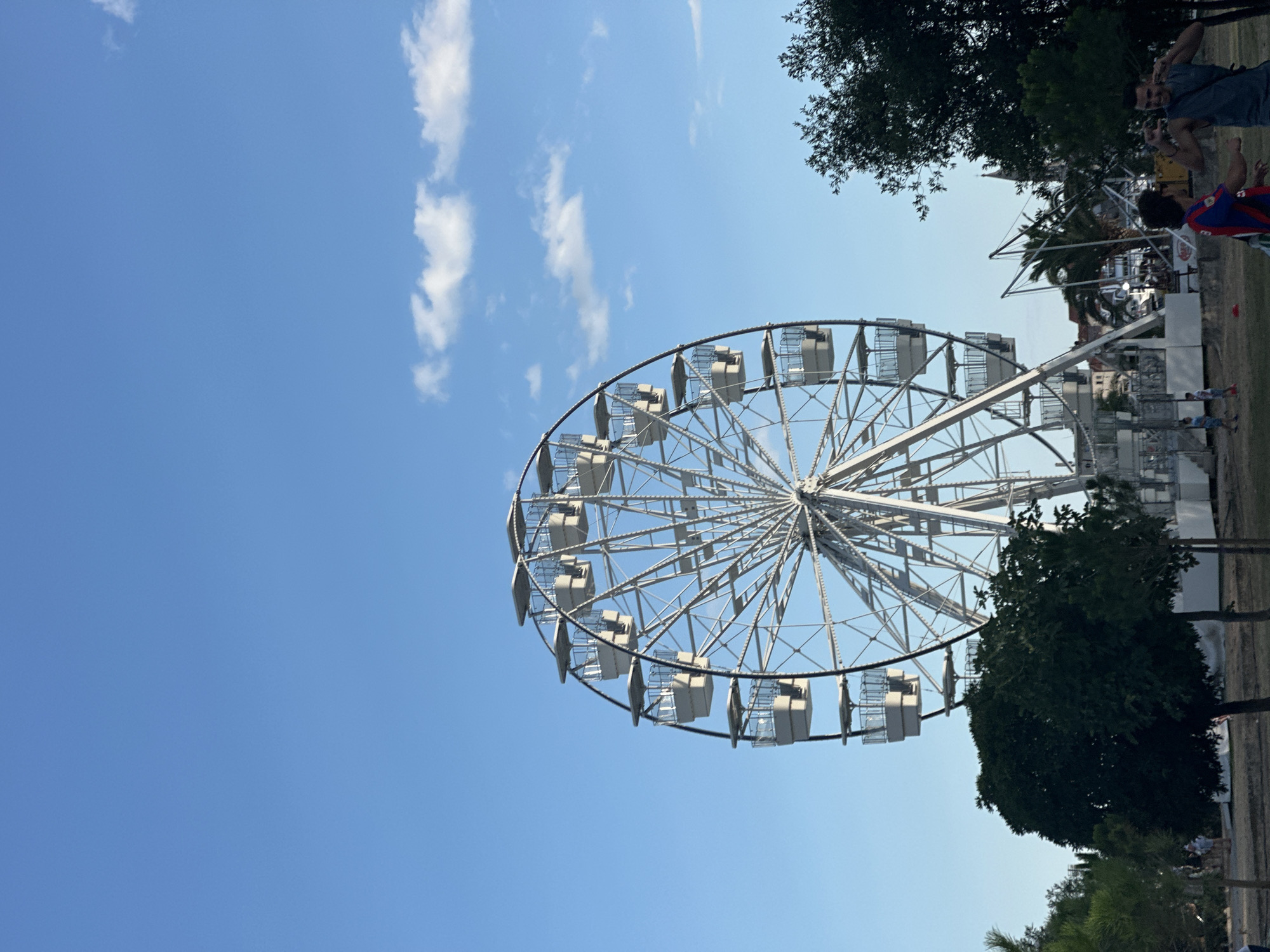 A large, white Ferris wheel stands prominently against a clear blue sky with a few scattered white clouds. The Ferris wheel has multiple white cabins and intricate metal supports. In the foreground, there are various trees with dark green foliage. To the right of the Ferris wheel, in the mid-ground, a few people are visible. One person is wearing a dark grey tank top and appears to be making a gesture with their hands. Another person is wearing a red, blue, and yellow striped shirt. A third person is wearing a blue shirt. The environment appears to be an outdoor park or amusement area, possibly in Budva, Montenegro, given the context. There are some buildings visible in the background behind the trees and the Ferris wheel.