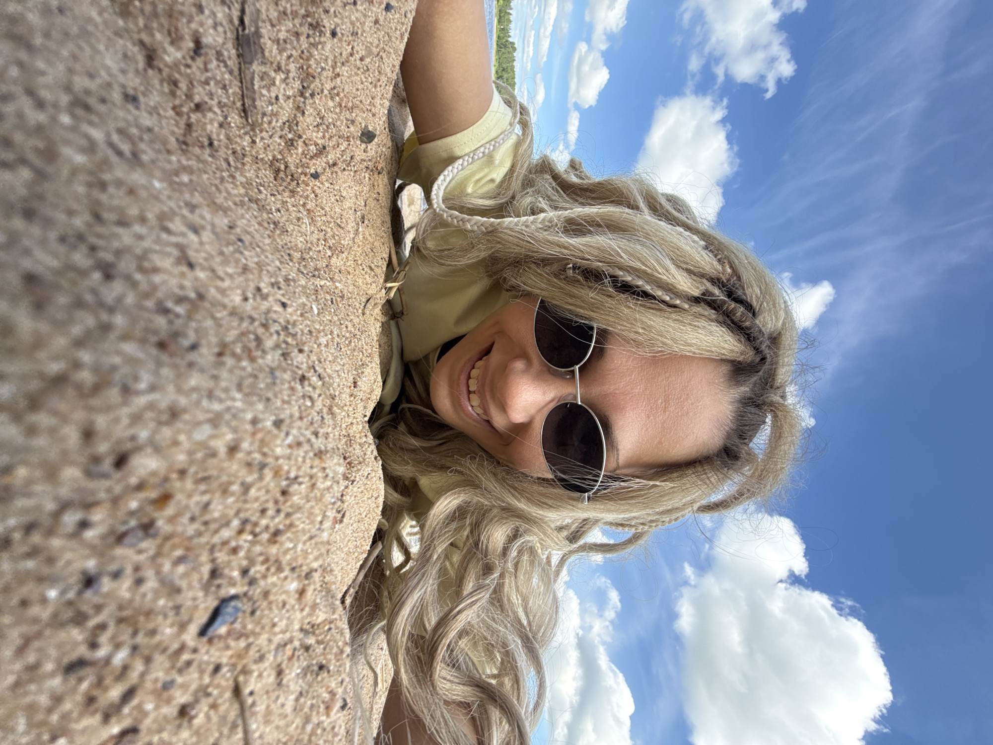 The image captures a smiling person, presumably a woman, with long, light blonde hair featuring braided strands, wearing dark sunglasses and a light yellow t-shirt. She is looking directly at the camera with a wide, happy smile.
The photo is taken from a low, upward-looking angle, with the subject positioned as if lying down or leaning against an incline. The image itself is oriented sideways, giving the impression that she is looking down or to the side.
The left side of the frame features a close-up, slightly out-of-focus view of sandy ground, suggesting a beach or lakeside environment. The right side of the frame reveals a clear, bright blue sky dotted with fluffy white clouds. In the background, beyond the person's head, a body of water (likely a lake or large river) with a green treeline on the opposite shore is visible.
The scene depicts a relaxed outdoor moment, likely a selfie, on a sunny day with pleasant weather. The person's expression conveys happiness and enjoyment. The location cues, combined with the provided context, suggest this scene takes place in Rusanovo, Russia, possibly by a natural water body. There is no visible text in the image.