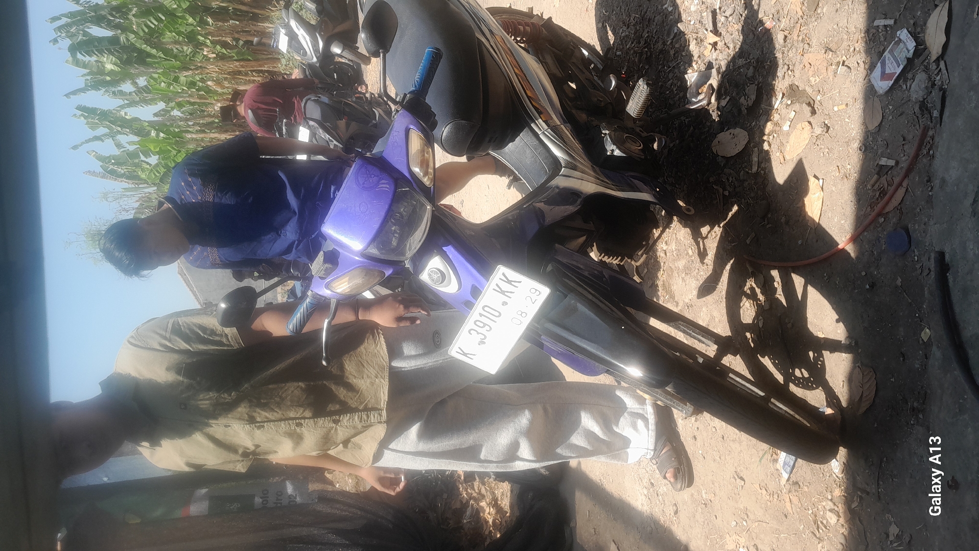 In the city of Hadipolo, Indonesia, a sunny day is captured with an outdoor scene featuring several individuals and motorcycles. In the foreground, a purple and black motorcycle with the license plate K 3910 KK is prominently displayed. A person wearing light-colored sweatpants and a khaki shirt stands to the left of the motorcycle, partially obscuring it. To the right and slightly behind, another individual in a blue shirt is visible, with a person in a maroon shirt further back. Banana trees line the background, suggesting a rural or semi-rural setting. The ground is unpaved, covered with dirt and scattered debris, including cigarette butts and fallen leaves. The lighting indicates it is daytime, with distinct shadows cast by the people and motorcycles.